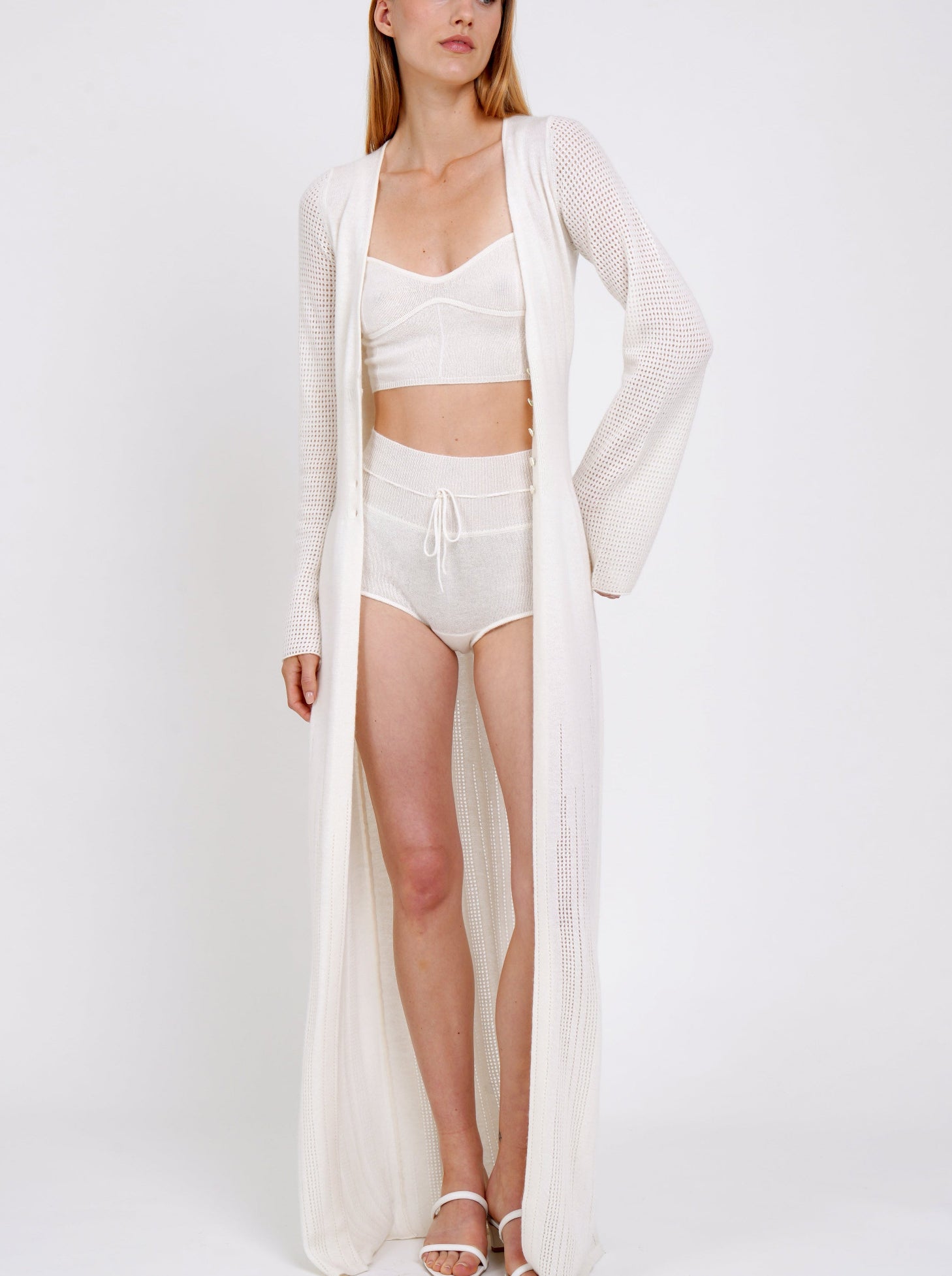 standing model wears long white knit cardigan over white cashmere shorts with drawstring waist and matching white knit cropped bra top.