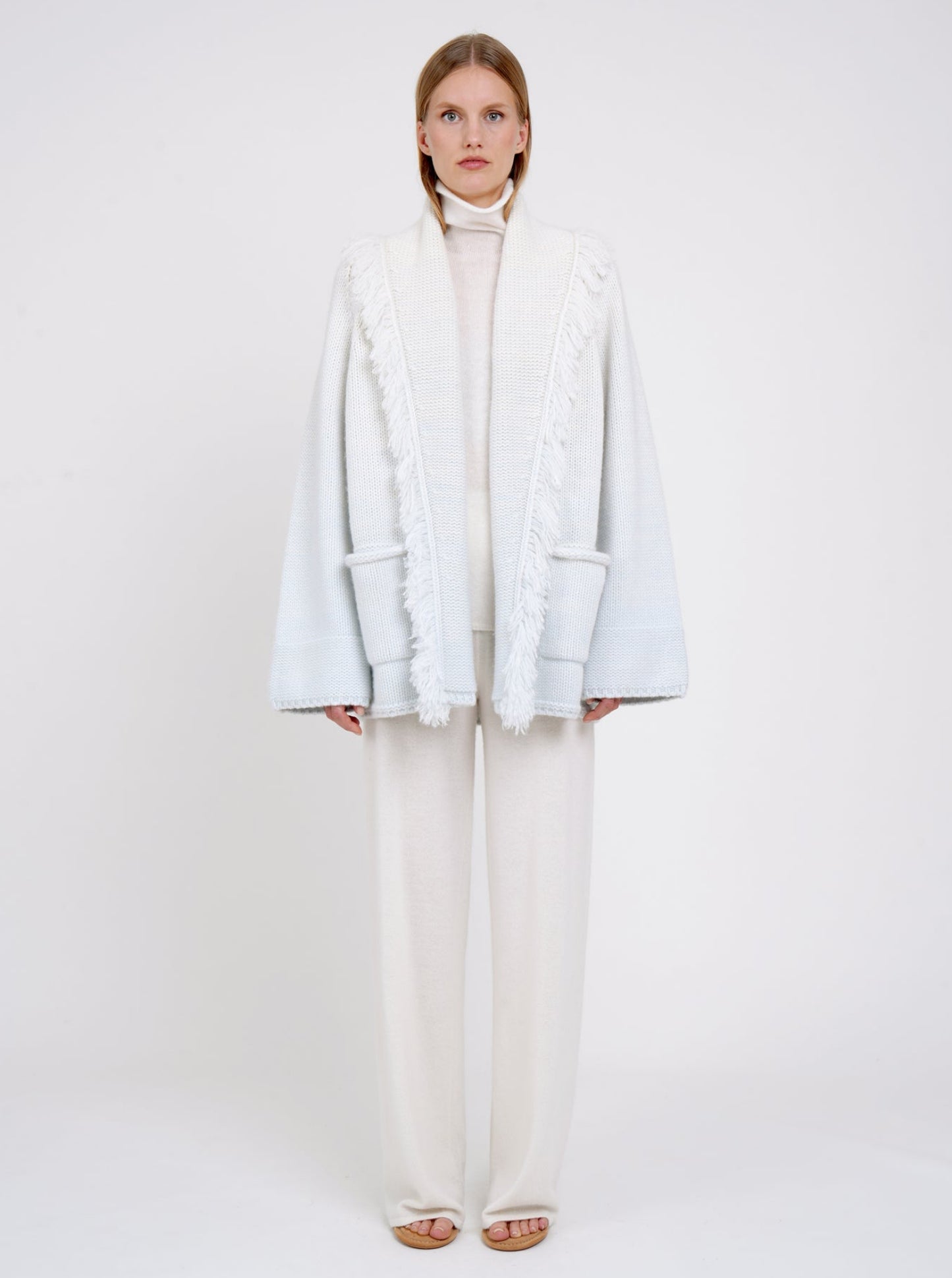 Model wears chunky knit cashmere jacket in a blend of two colours, natural white and very pale blue. Worn with white cashmere turtle neck and white wide leg pants.