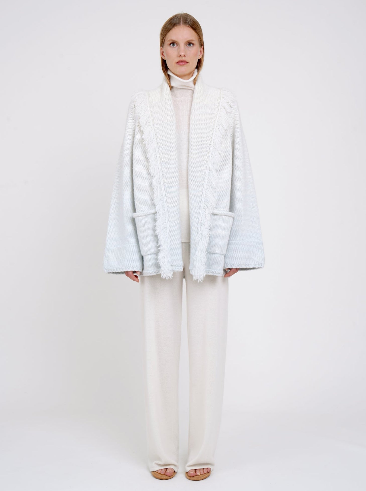 Model wears chunky knit cashmere jacket in a blend of two colours, natural white and very pale blue. Worn with white cashmere turtle neck and white wide leg pants.