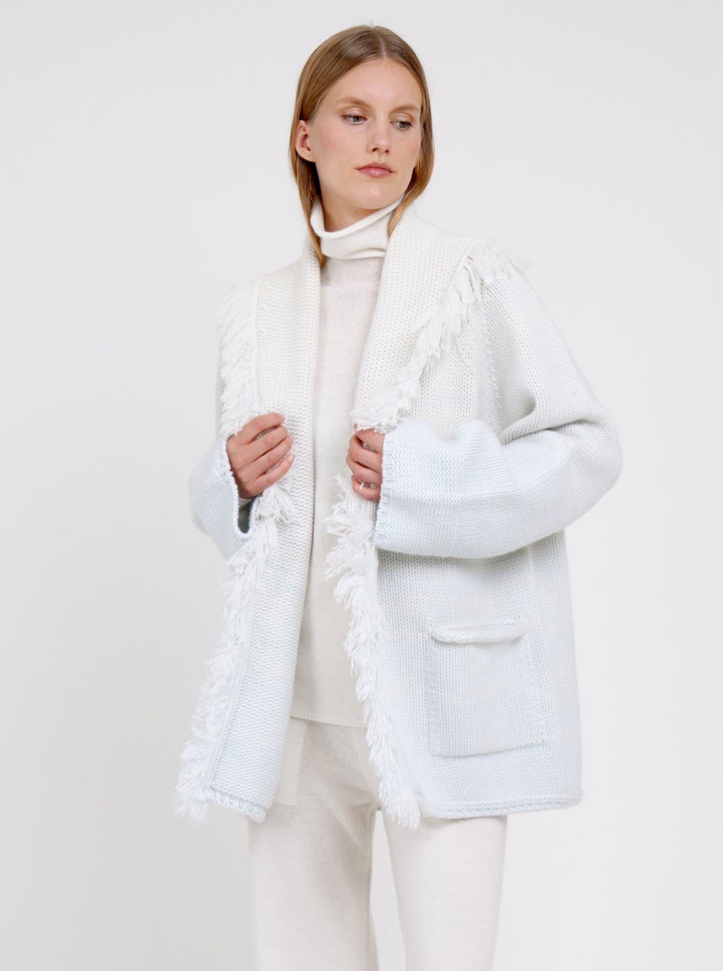 Close up image model wearing chunky knit cashmere jacket in a blend of two colours, natural white and very pale blue, with deep shawl collar with fringe trim and patch pockets.