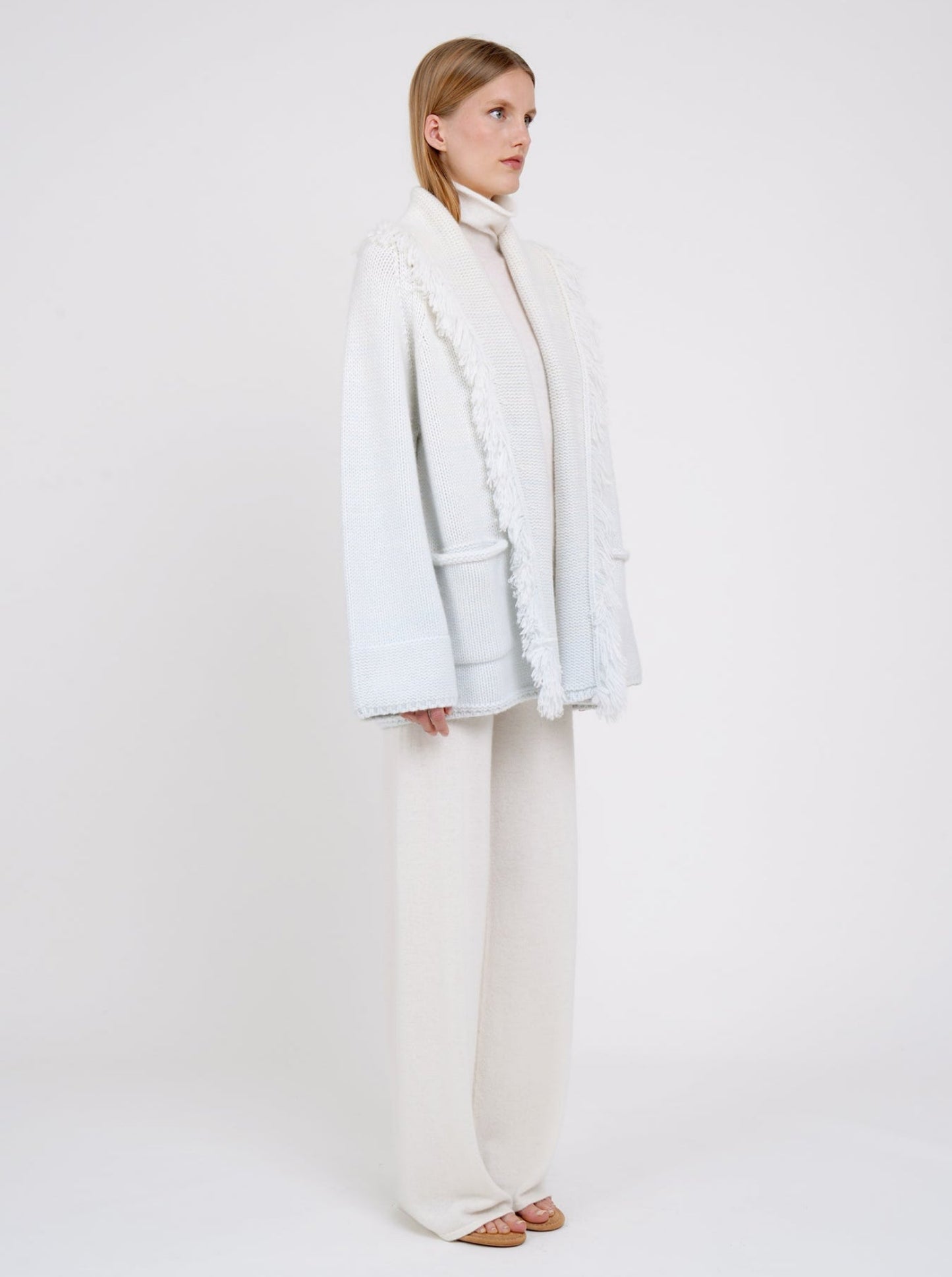 Side view model wearing chunky knit cashmere jacket in a blend of two colours, natural white and very pale blue. Worn with white cashmere turtle neck and white cashmere wide leg pants.
