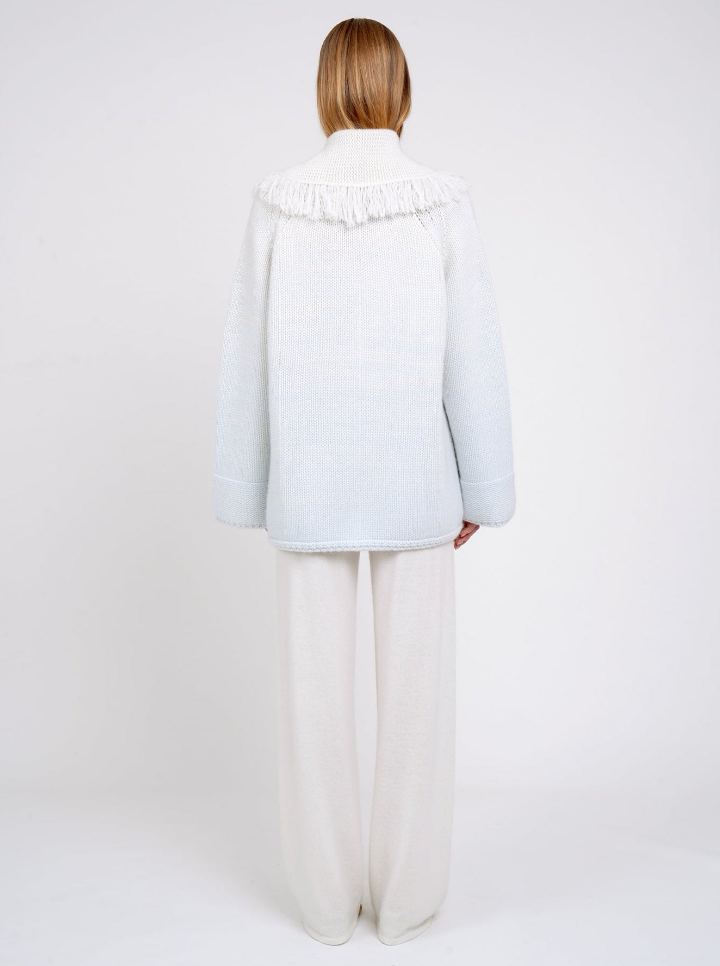 Back view model wearing chunky knit cashmere jacket with deep shawl collar, in two mixed colours, natural white and very pale blue. Worn with white wide leg pants.