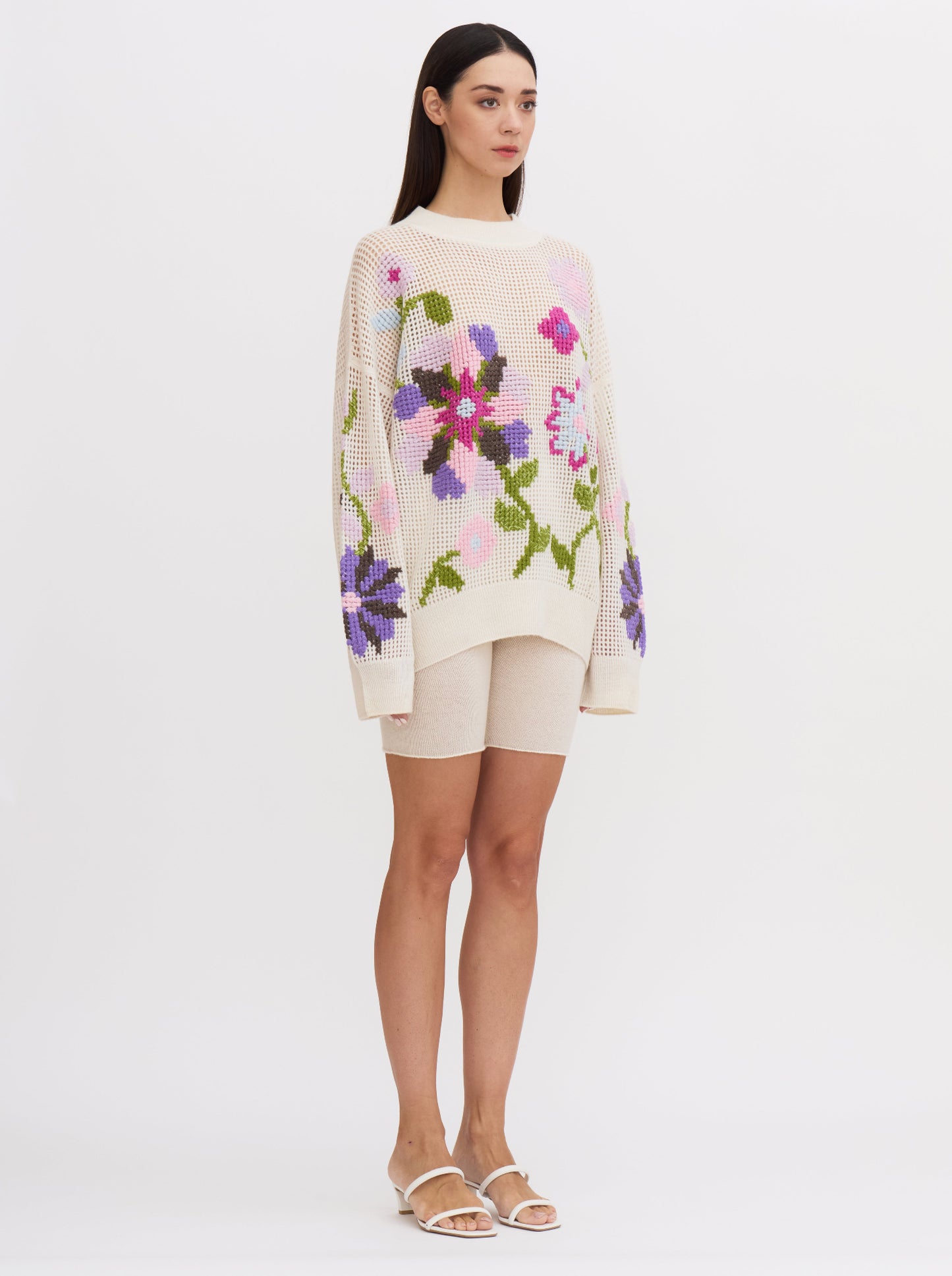 Model wears white net-knit cashmere oversize jumper with round neck and embellished with hand-embroidered cross-stitch large floral pattern in multi-coloured cashmere yarns; worn with white cashmere cycling shorts.