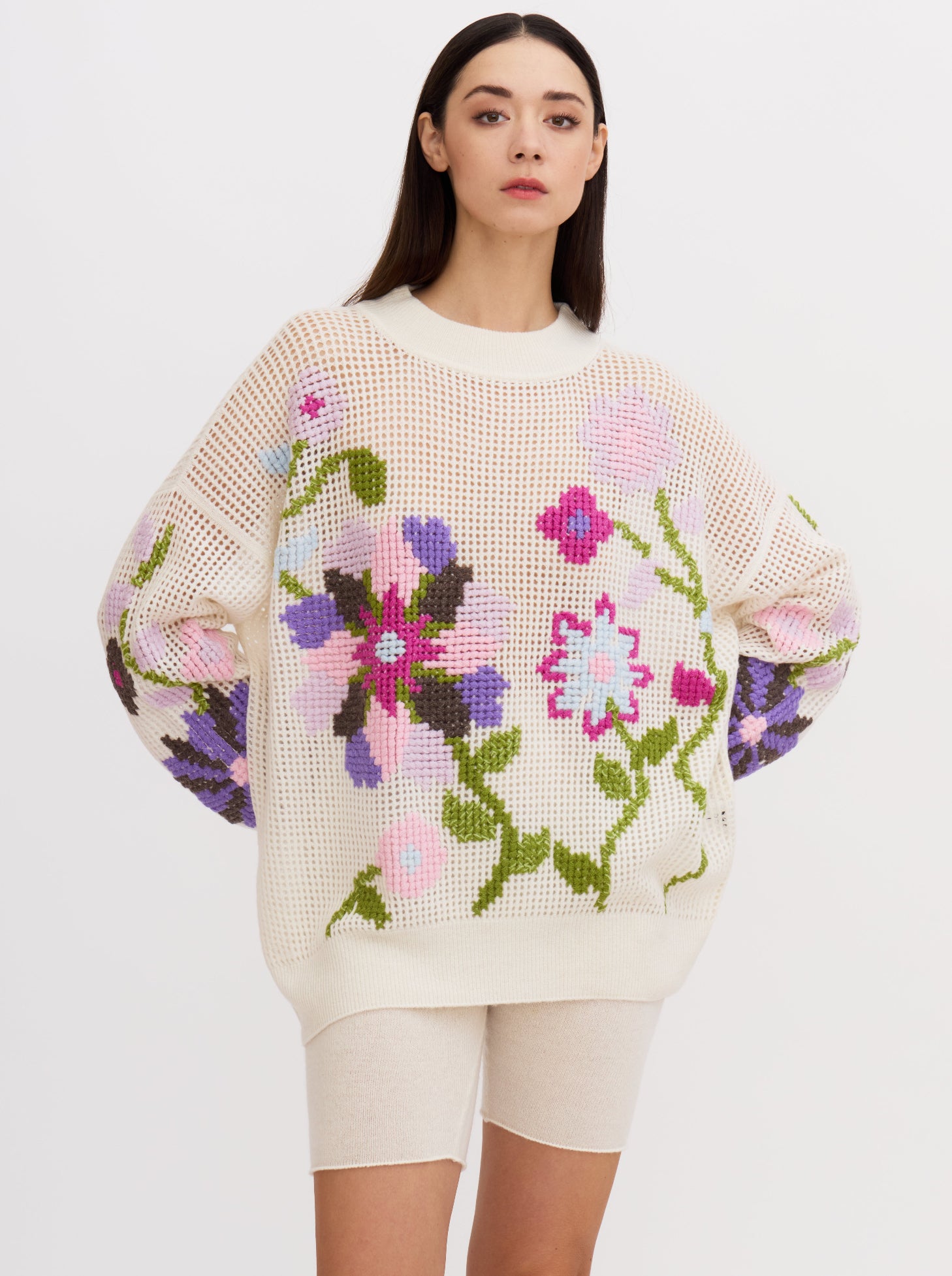 Close-up of model wearing white net-knit cashmere round neck jumper embellished with hand-embroidered cross-stitch large floral pattern in multi-coloured cashmere yarns; worn with white cashmere cycling shorts.