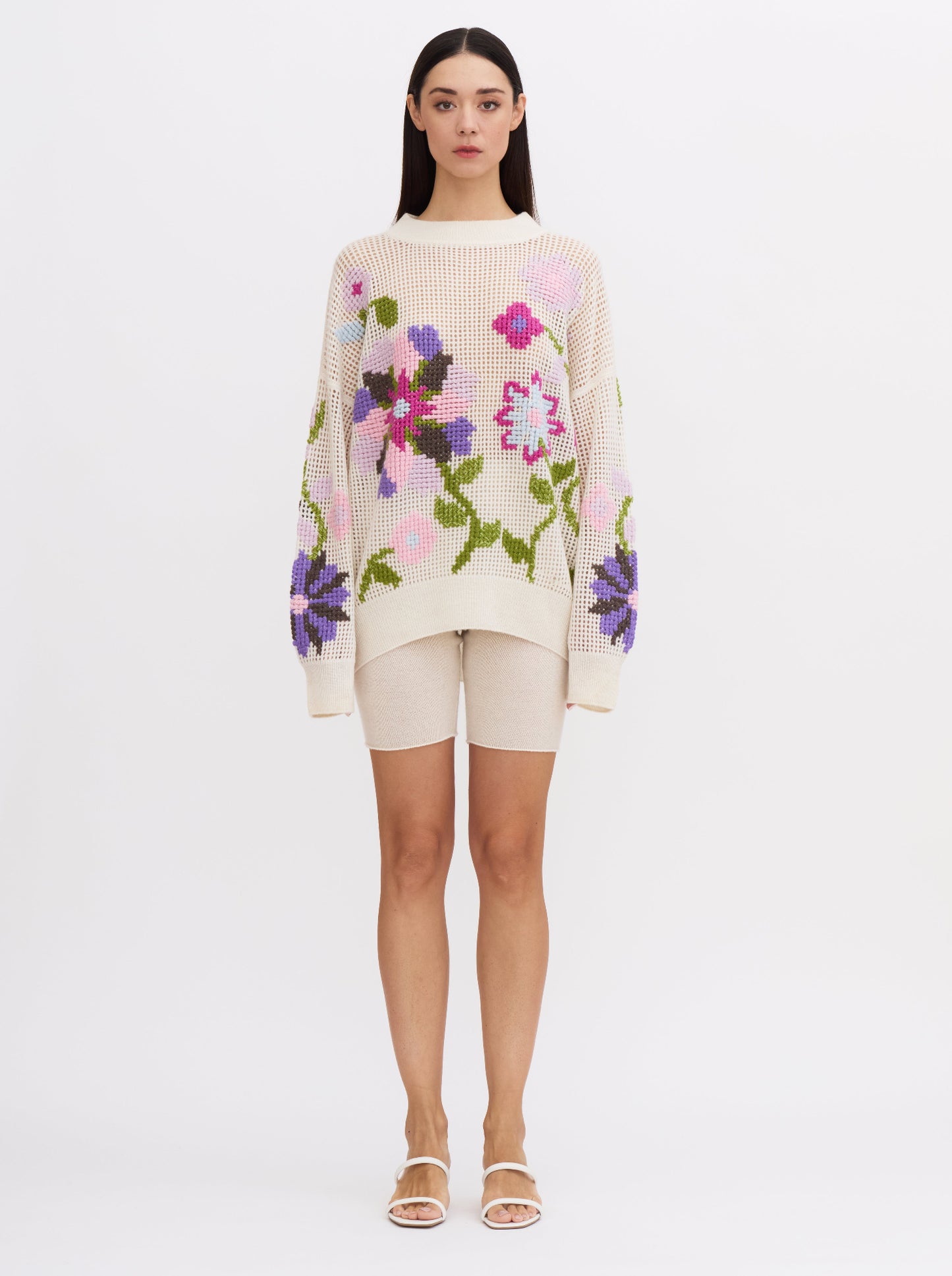 Front view of model wearing white net-knit cashmere round neck sweater with hand-embroidered cross-stitch large floral pattern in multi-coloured cashmere yarns; worn with white cashmere cycling shorts.