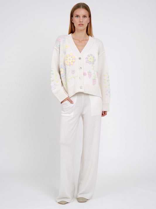 Model wearing a white cashmere V neck cardigan with mother-of-pearl buttons and pastel colour floral embroidery worn with white cashmere pants.