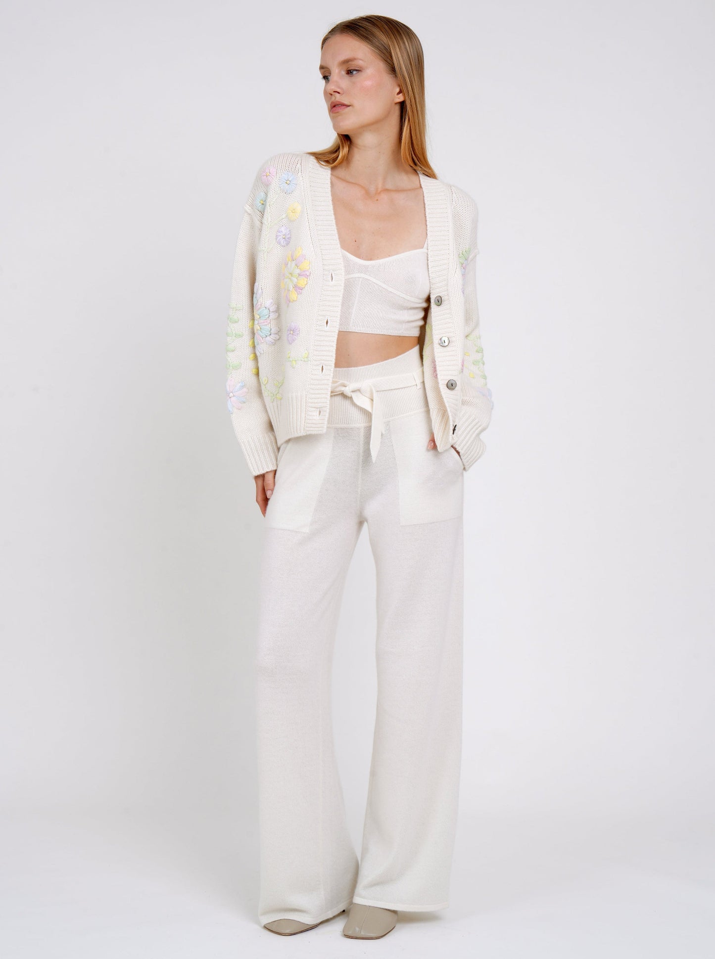 Model wearing a white cashmere V neck cardigan with mother-of-pearl buttons and pastel colour floral embroidery; worn undone showing white cashmere bralette underneath and cashmere belted pants.