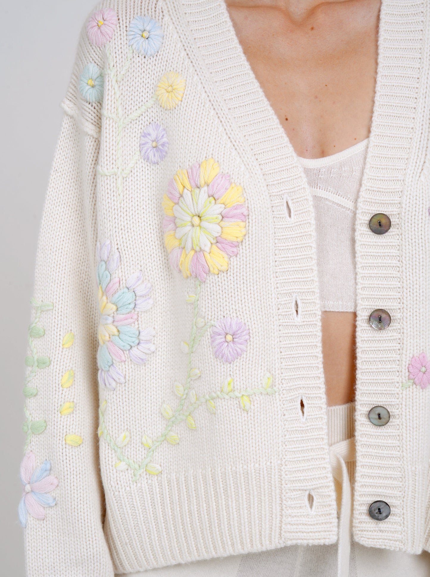 Cropped detail of model wearing a white cashmere V neck cardigan with mother-of-pearl buttons and pastel colour floral hand embroidery.