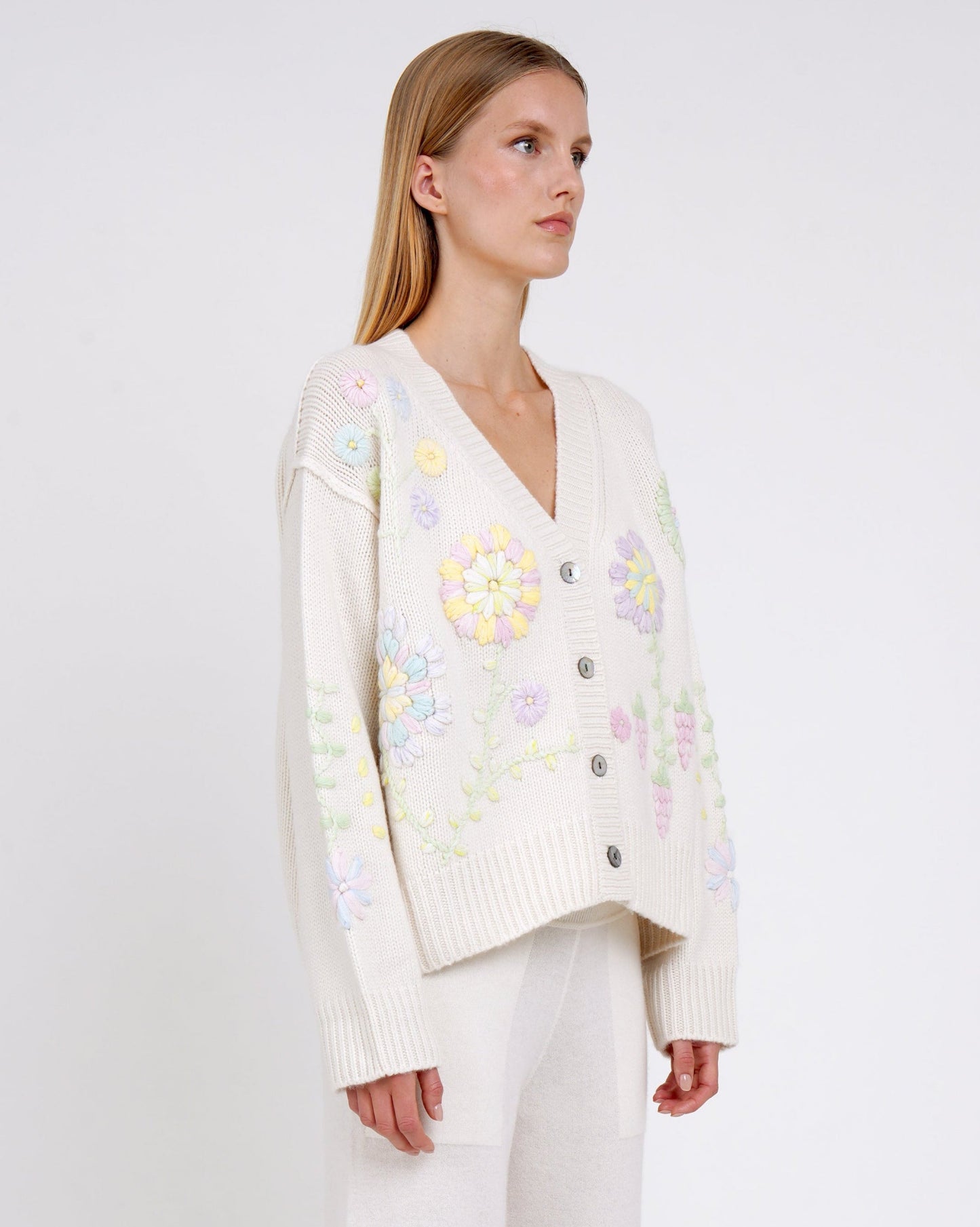 Close up of model wearing a white cashmere V neck cardigan with mother-of-pearl buttons and pastel colour floral embroidery worn with white cashmere pants.