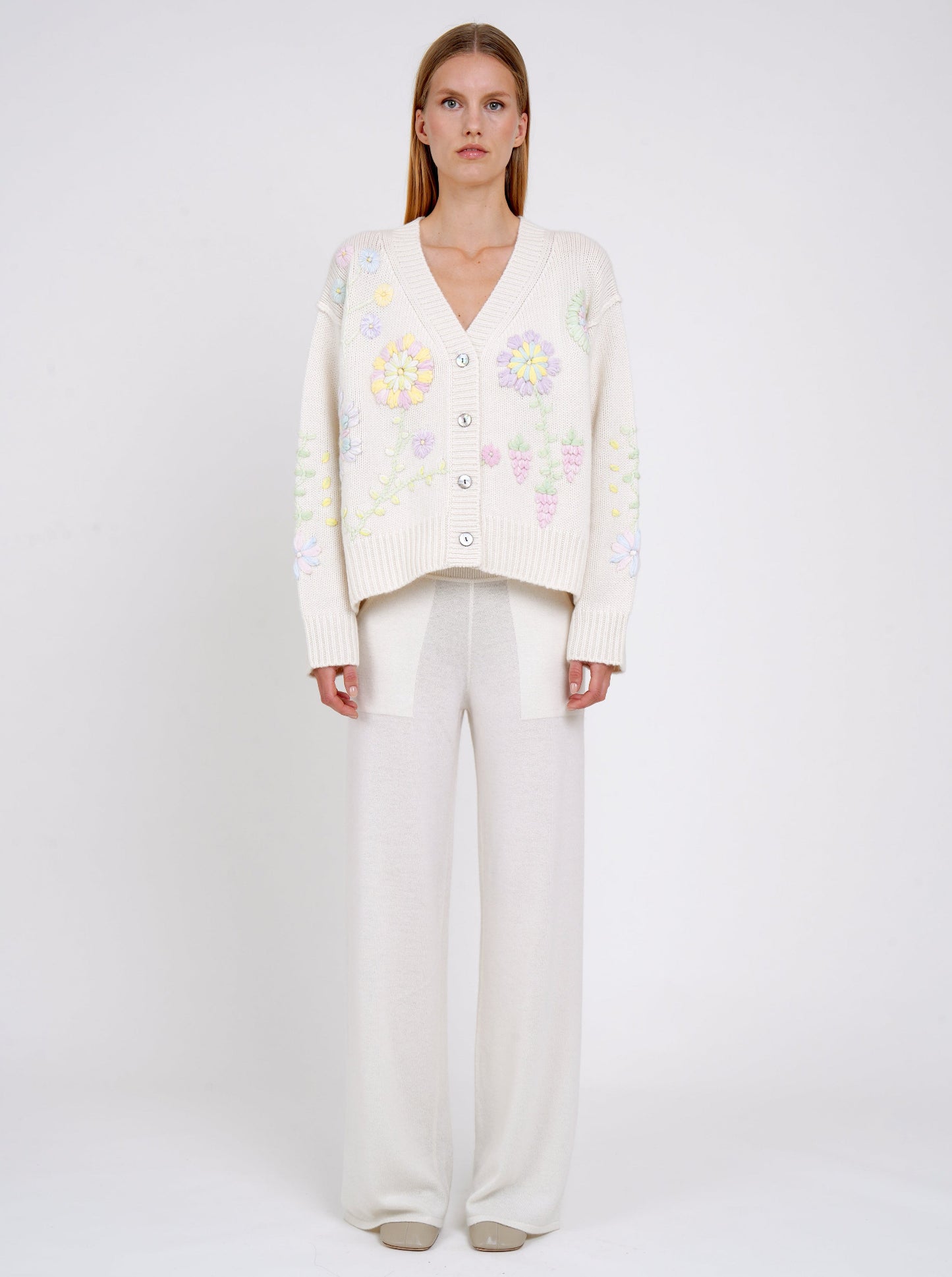 Front view model in white cashmere V neck cardigan with mother-of-pearl buttons and pastel colour floral hand embroidery worn with white cashmere wide-leg pants.