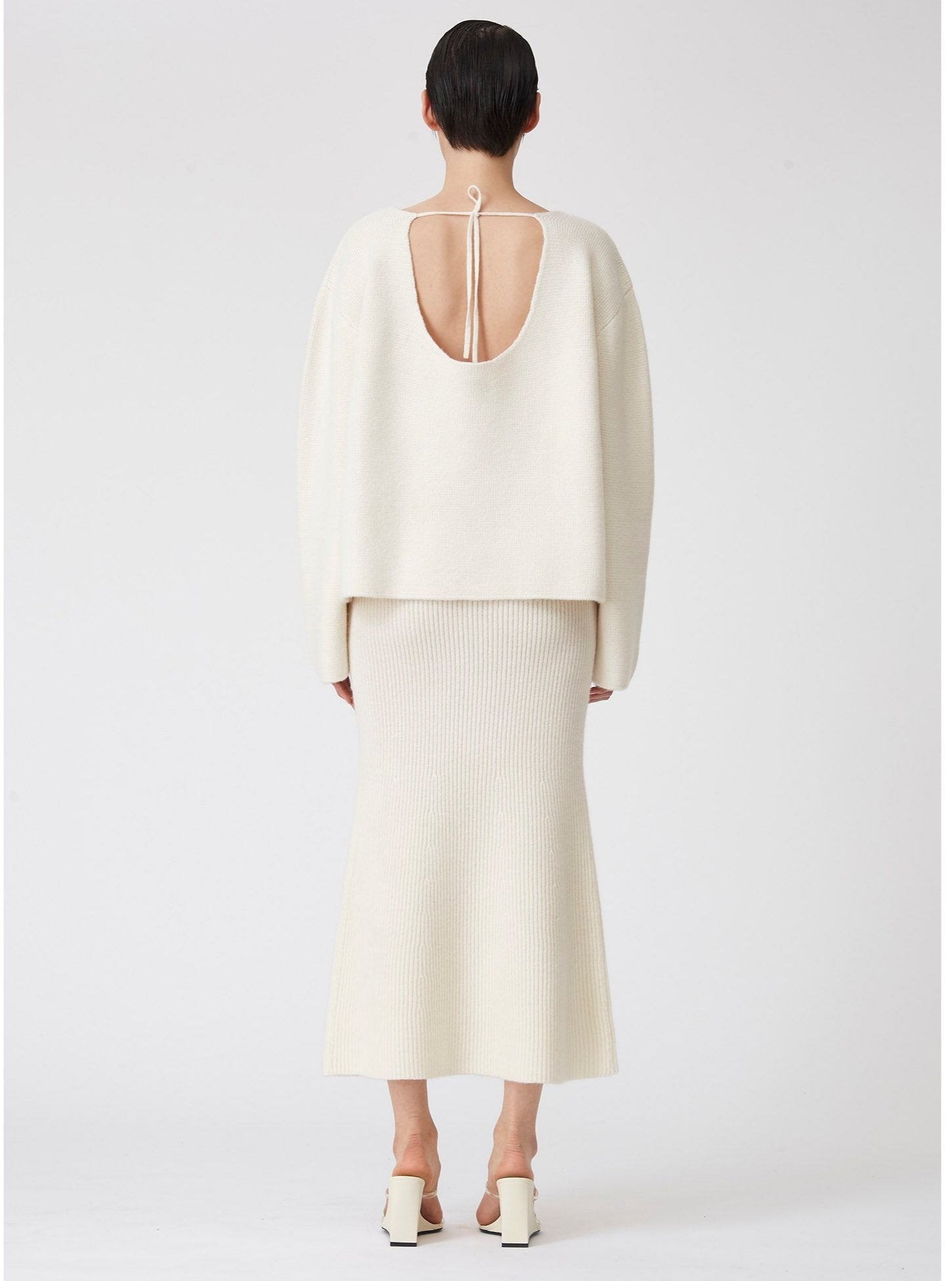 Back view of modal wearing white ribbed knit flared midi skirt with low plunge back white cashmere jumper.