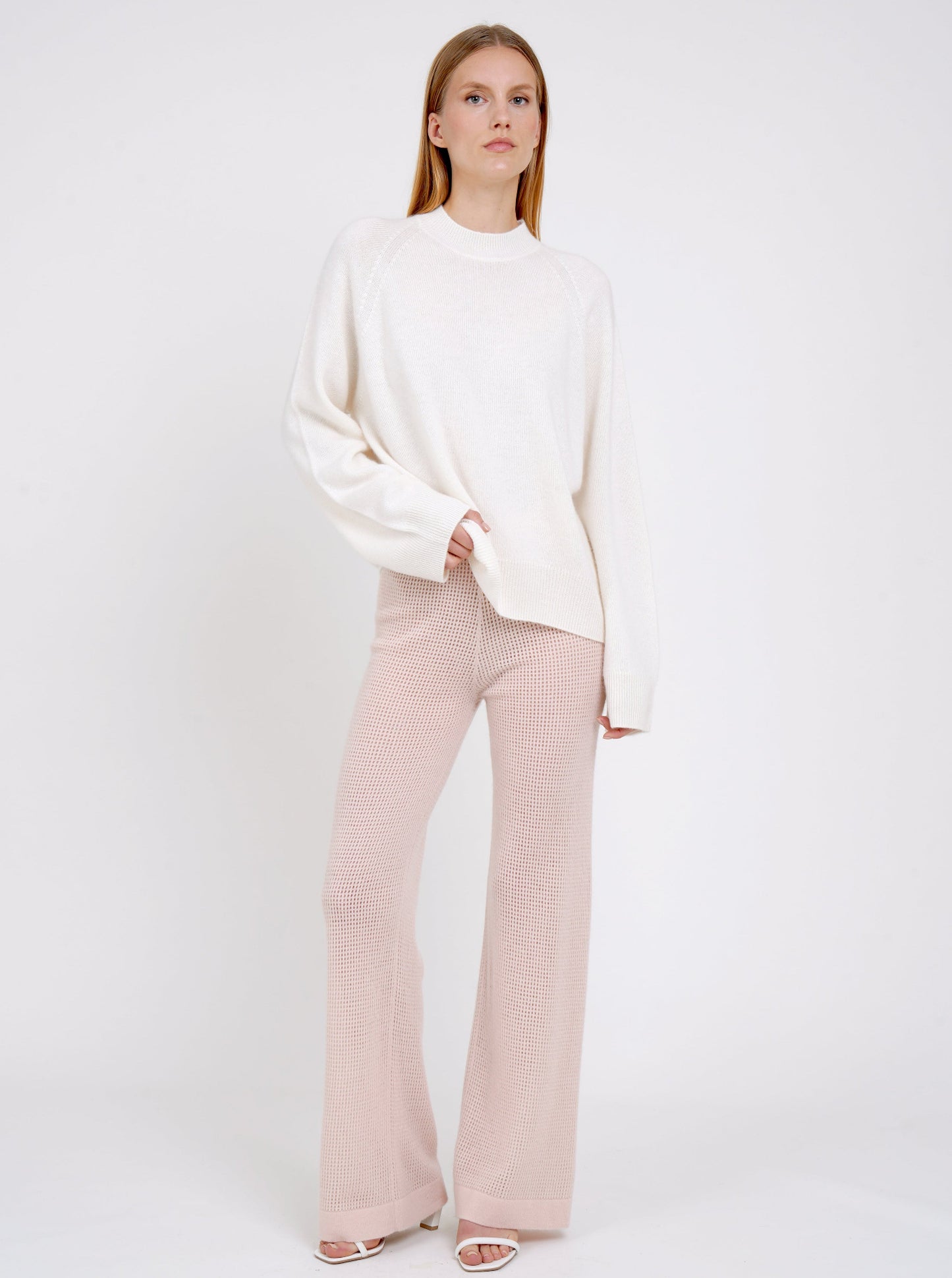 Standing model in white cashmere jumper with round neck, long sleeves and pale peach pink knit cashmere pants.