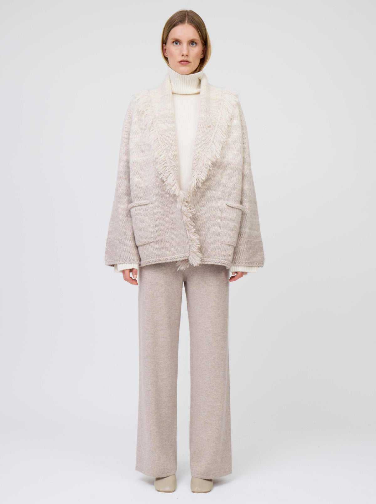 Model wears  gradient colour mix knitted cashmere jacket in white and pale beige over white cashmere roll neck sweater and beige wide leg cashmere pants.