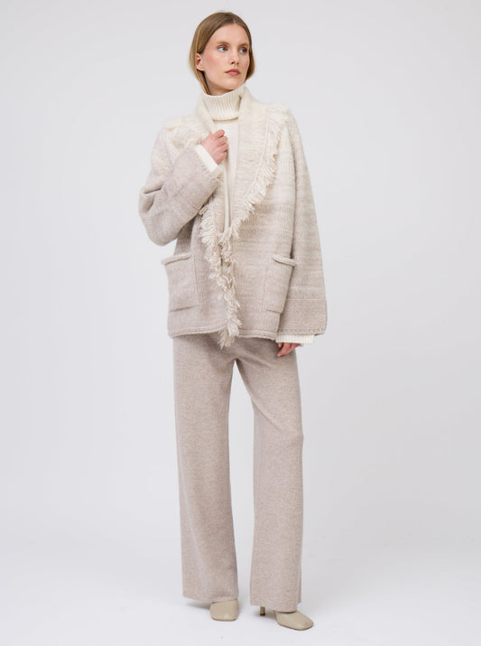 Model wears  gradient colour mix knitted cashmere jacket with fringed shawl collar in white and pale beige over white cashmere roll neck sweater and beige wide leg cashmere pants.