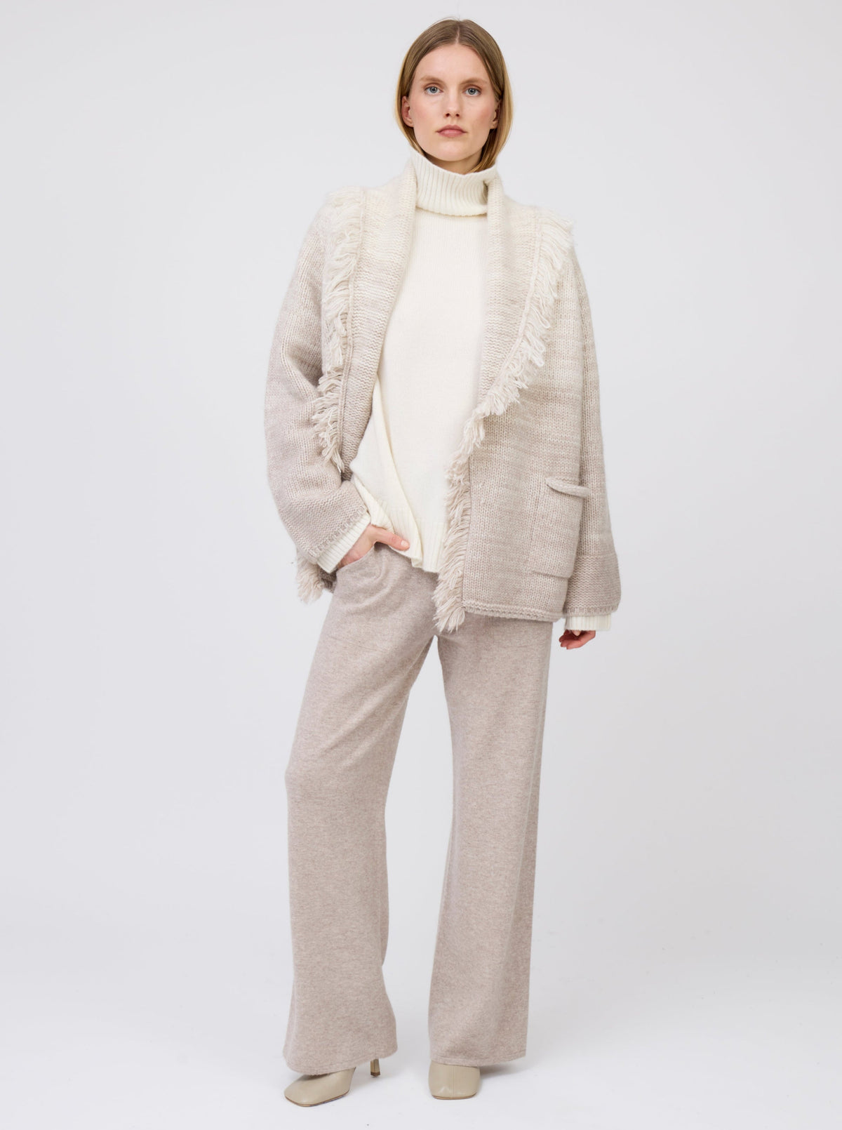 Model in relaxed pose, wears  two colour mix knitted cashmere jacket with shawl collar and pockets in white and beige over white cashmere roll neck sweater and beige wide leg cashmere pants.