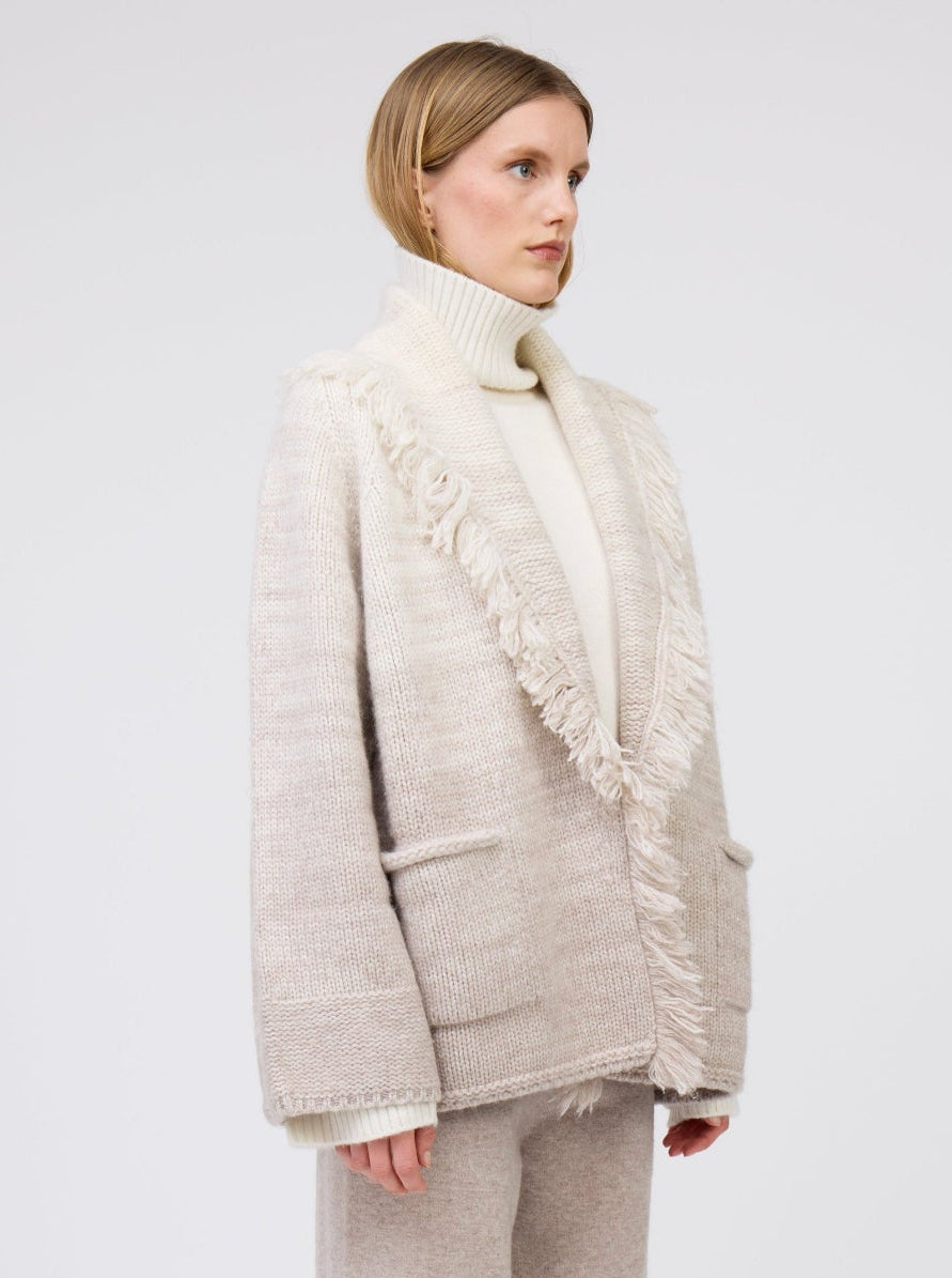 Model wears 2 colour mix knitted cashmere jacket with fringed shawl collar in white and beige over white cashmere roll neck sweater and beige wide leg cashmere pants.
