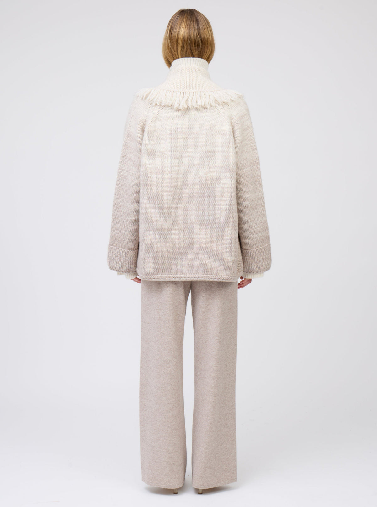 Back view of model wearing  gradient colour mix knitted cashmere jacket with deep shawl collar in white and pale beige over beige wide leg cashmere pants.