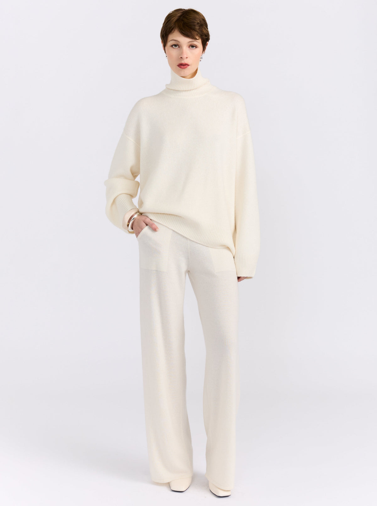 Front view of model in relaxed pose in white studio wearing white roll neck sweater with white knit wide leg pants and white high heels.
