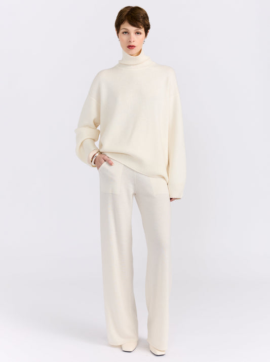 Front view of model in relaxed pose in white studio wearing white roll neck sweater with white knit wide leg pants and white high heels.