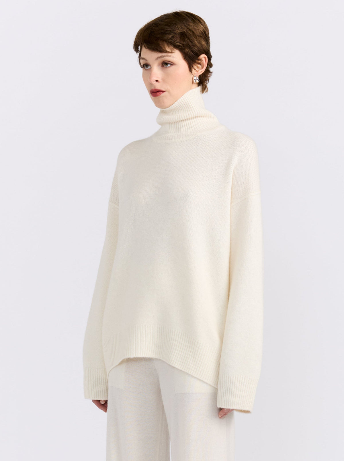 Close of side view of model in white studio wearing white roll neck sweater with white knit wide leg pants.