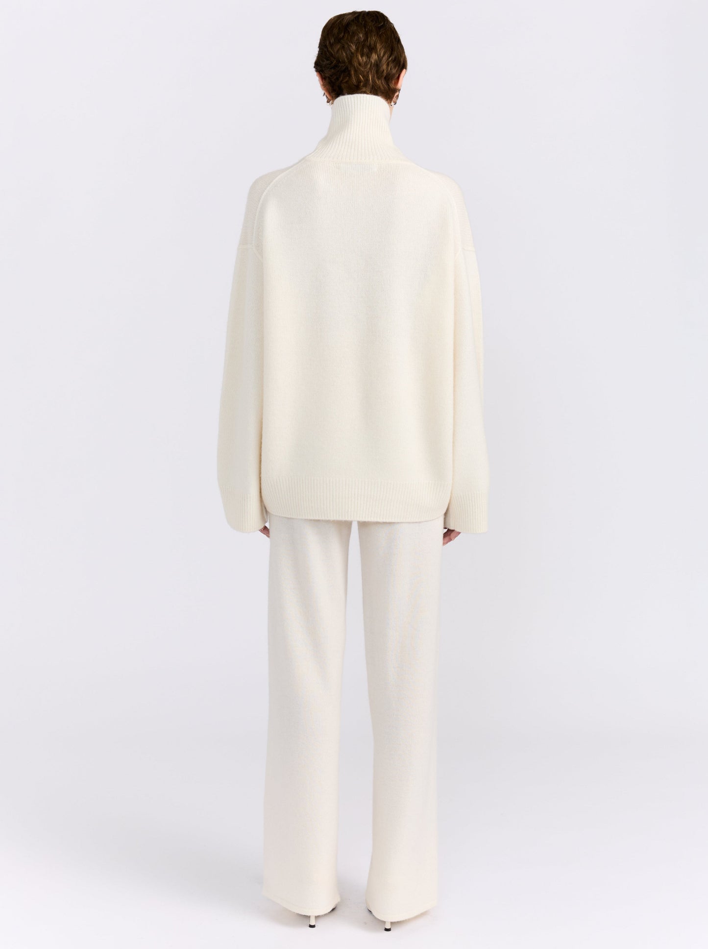 Back view of model standing in white studio wearing white roll neck sweater with white knit wide leg pants and white high heels.