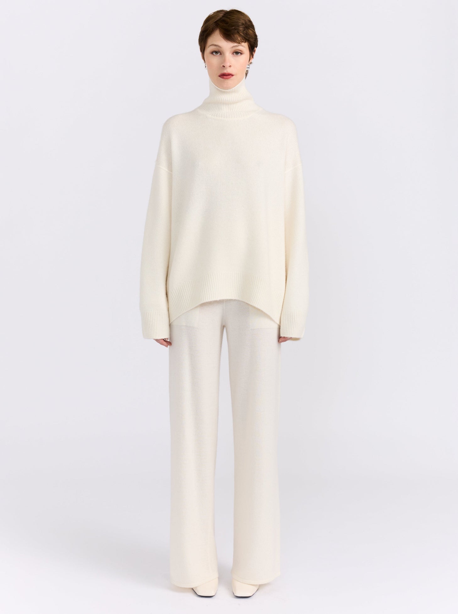 Front view of model standing straight in white studio wearing white roll neck sweater with white knit wide leg pants and white high heels.