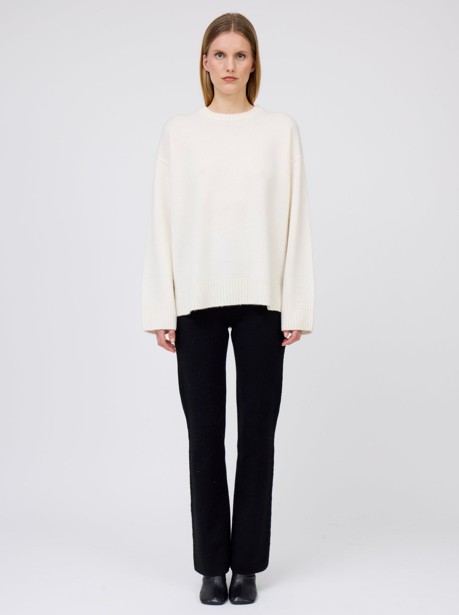 Front view of model wearing creamy white oversized round neck sweater with black suit pants and black boots.