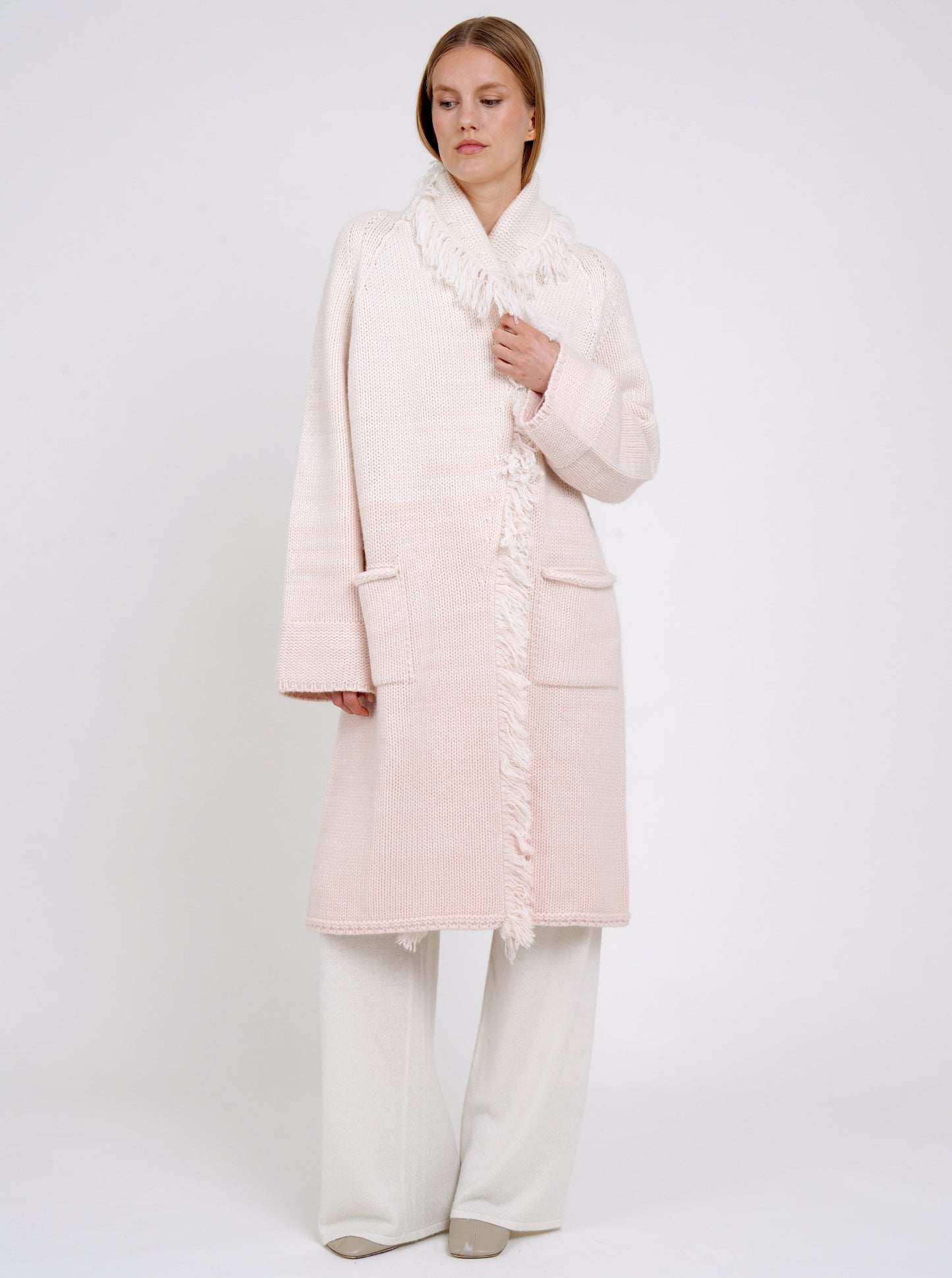 Model wears chunky knit loose coat in pale peach cashmere blended with white, with shawl collar, fringe trim, patch pockets, worn with white cashmere wide leg trousers.
