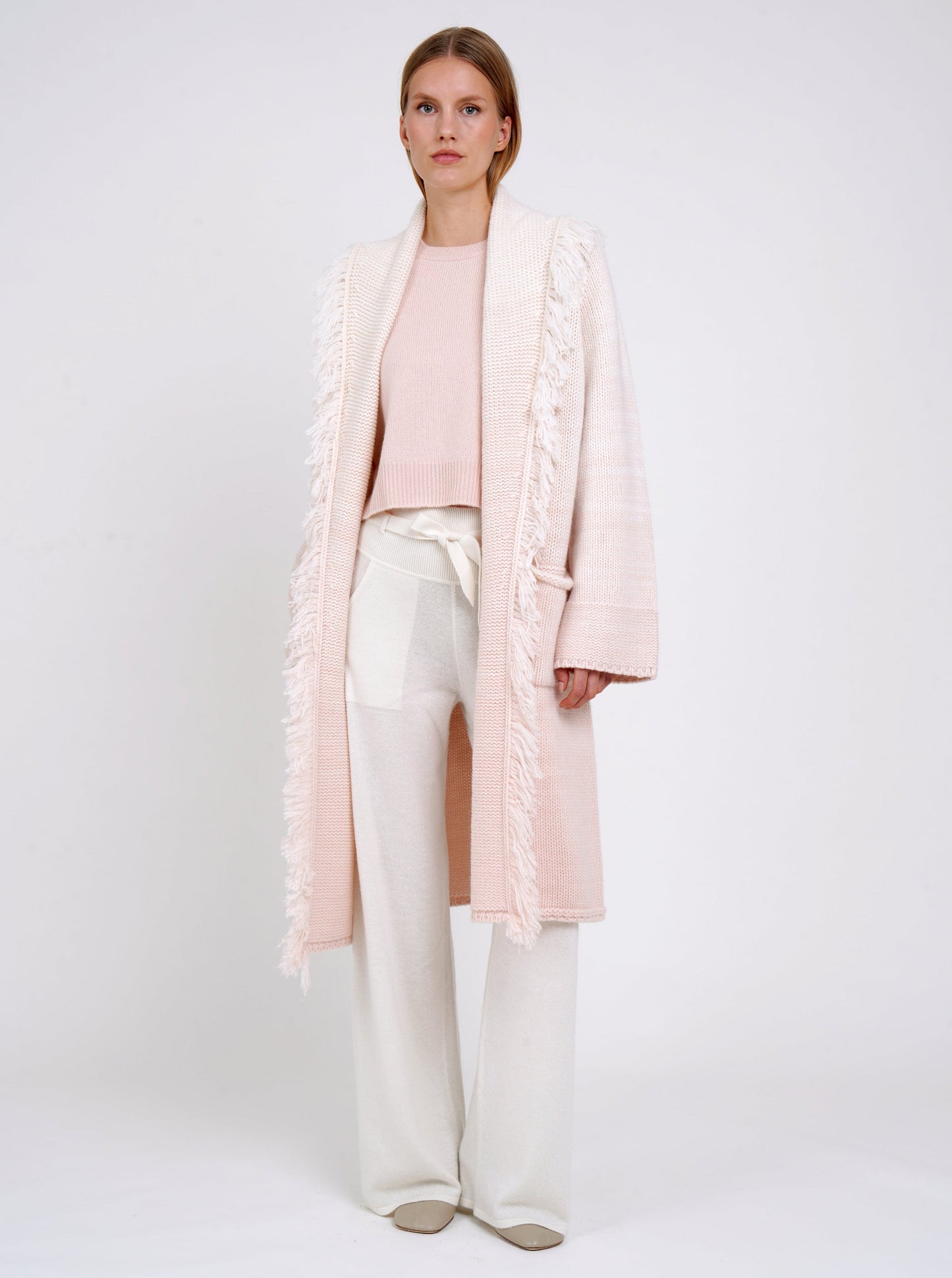 Model wears chunky knit loose coat in pale peach cashmere blended with white, with shawl collar, fringe trim, patch pockets, worn with white cashmere wide leg trousers.