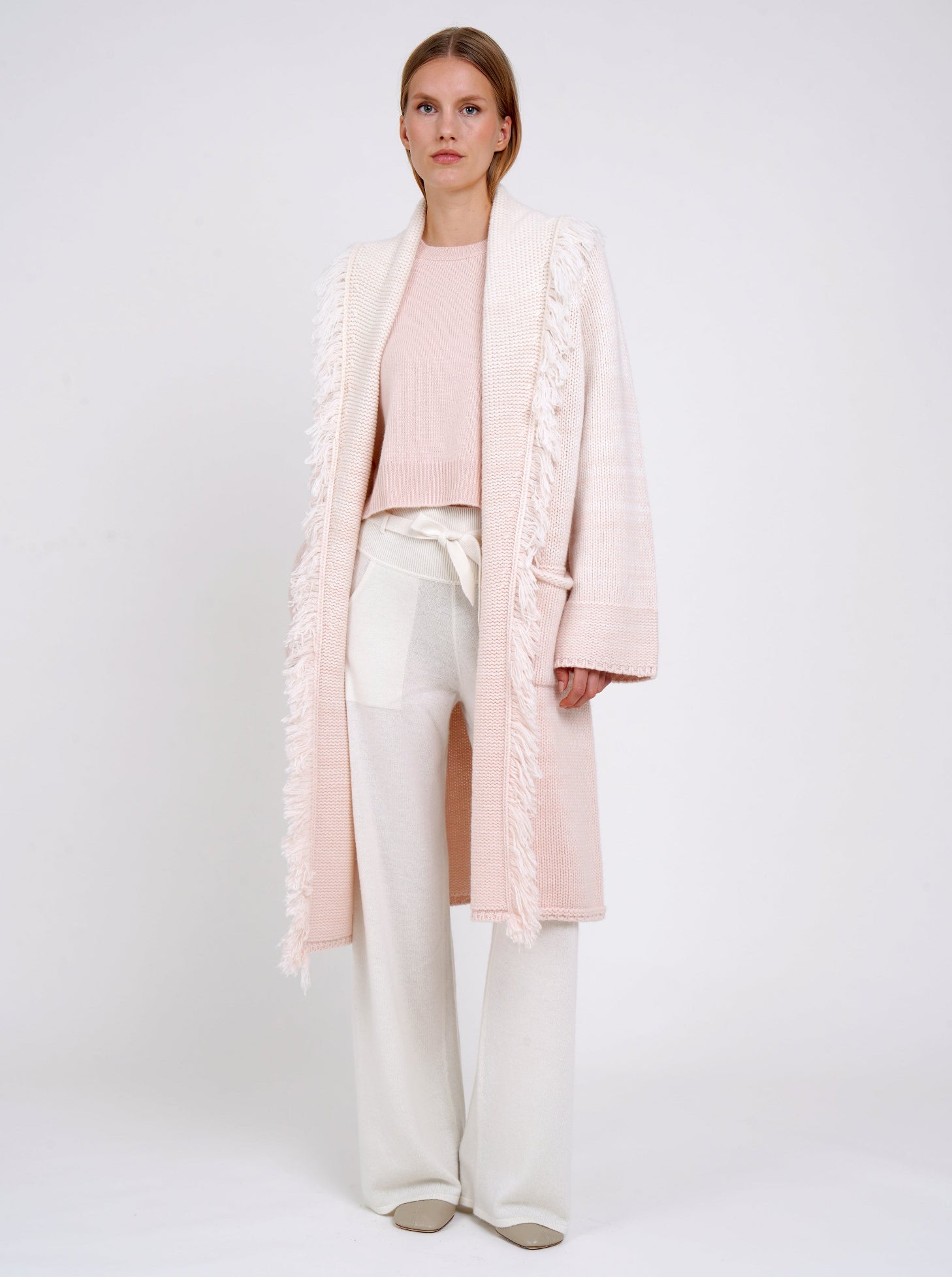 Model wears chunky knit loose coat in pale peach cashmere blended with white, with shawl collar, fringe trim, patch pockets, worn with white cashmere wide leg trousers.
