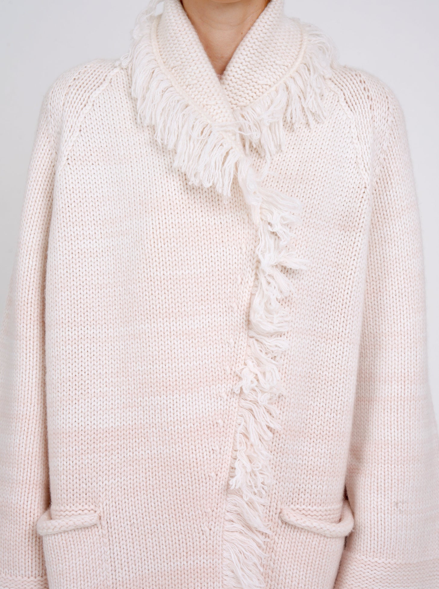 close-up showing chunky knit detail of coat in pale peach cashmere blended with white, with shawl collar, fringe trim, patch pockets.