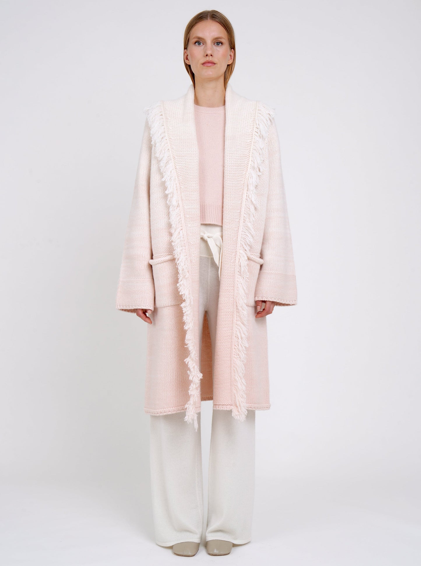 Front view of model in chunky knit loose coat in pale peach cashmere blended with white, with shawl collar, fringe trim, patch pockets, worn with white cashmere wide leg trousers.