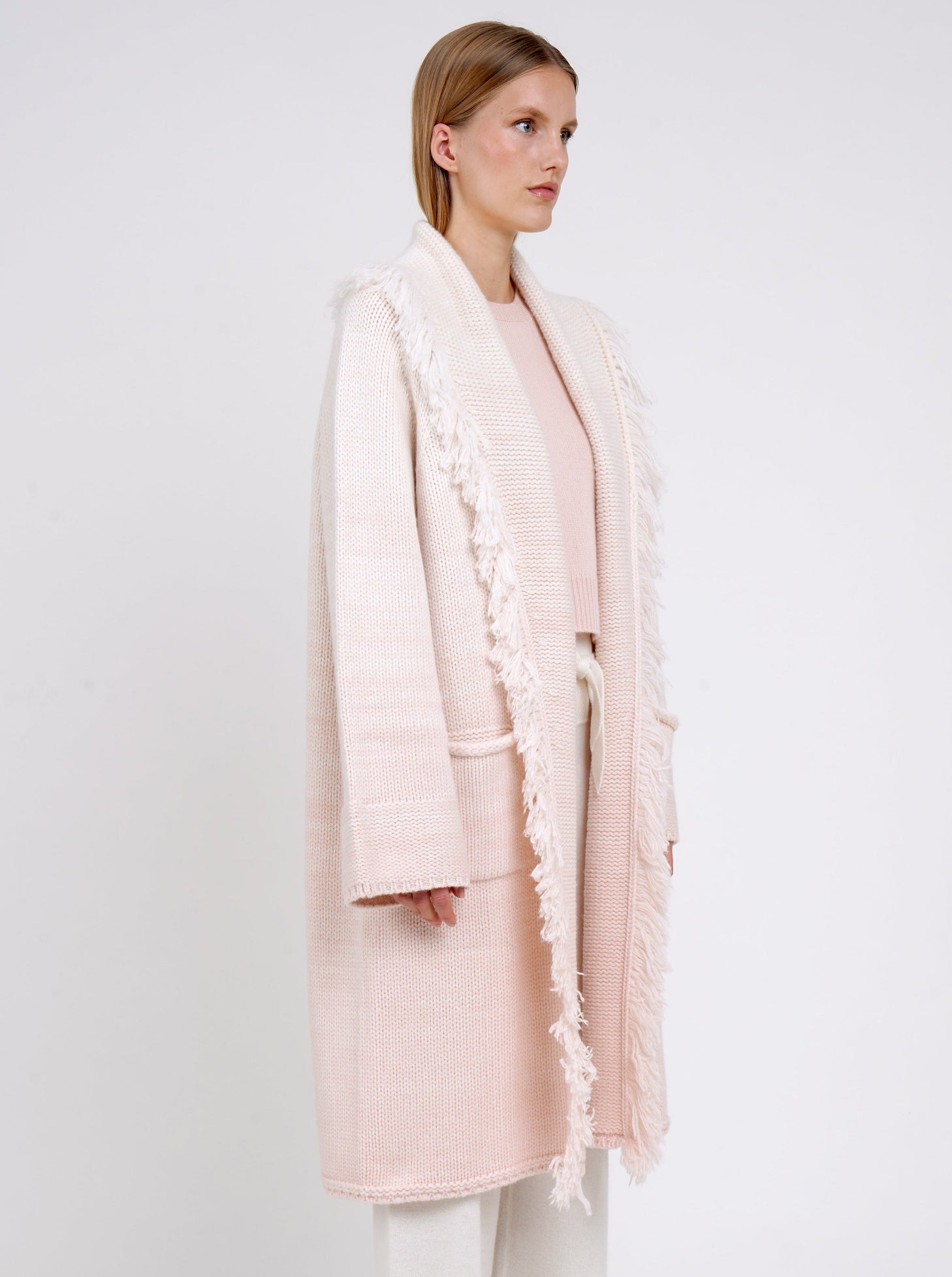 Side view model in chunky knit loose coat in pale peach cashmere blended with white, with shawl collar, fringe trim, patch pockets, with peach round neck jumper underneath.