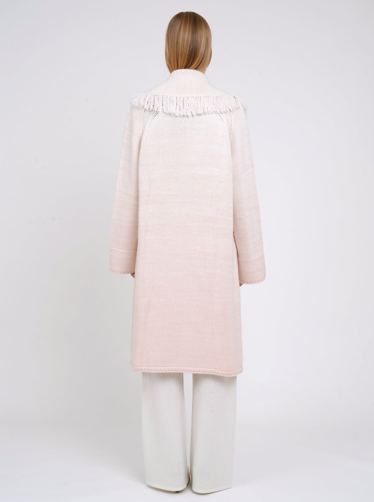 Back view of model in chunky knit loose coat in pale peach cashmere blended with white, with deep shawl collar, fringe trim, wide sleeves and worn with white cashmere wide leg trousers.