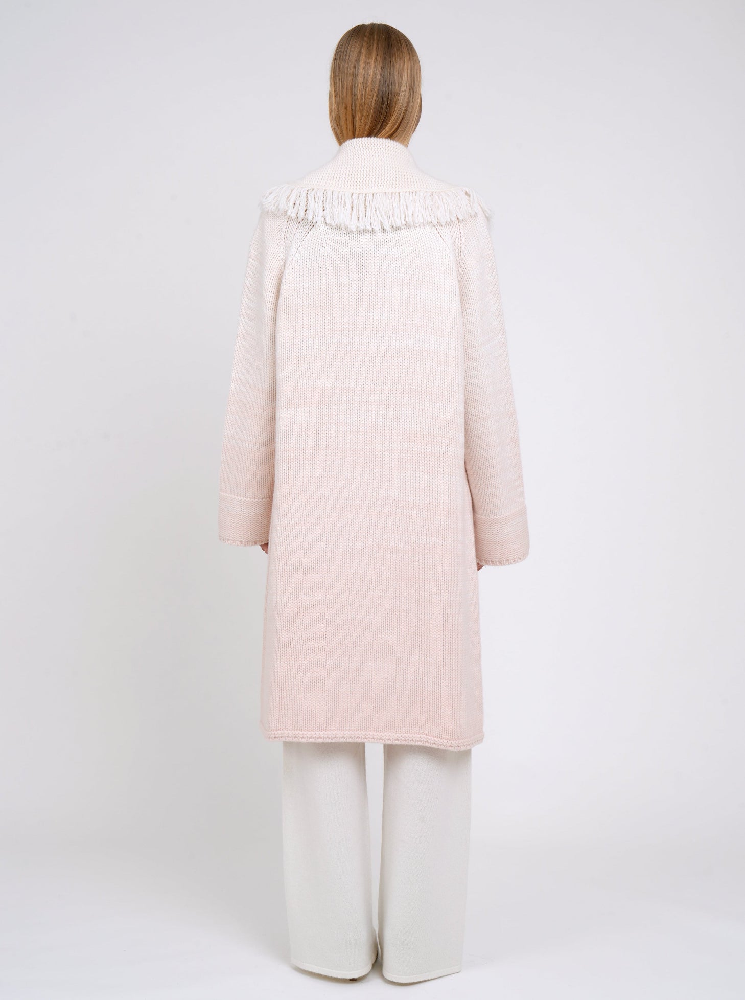 Back view of model in chunky knit loose coat in pale peach cashmere blended with white, with deep shawl collar, fringe trim, wide sleeves and worn with white cashmere wide leg trousers.