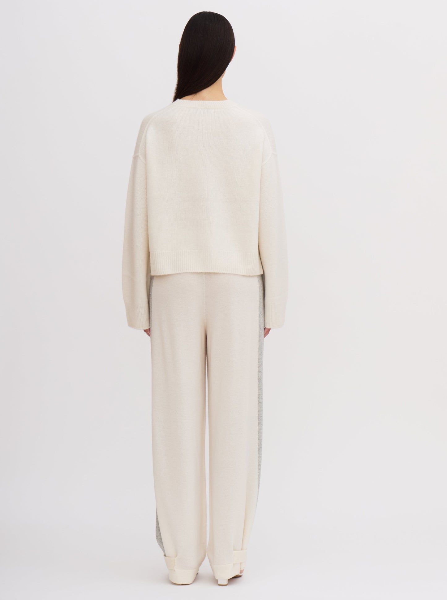 Back view of model wearing white wide leg pants with light grey side stripes and buttoned ankle strap detail; worn with matching white cashmere jumper.