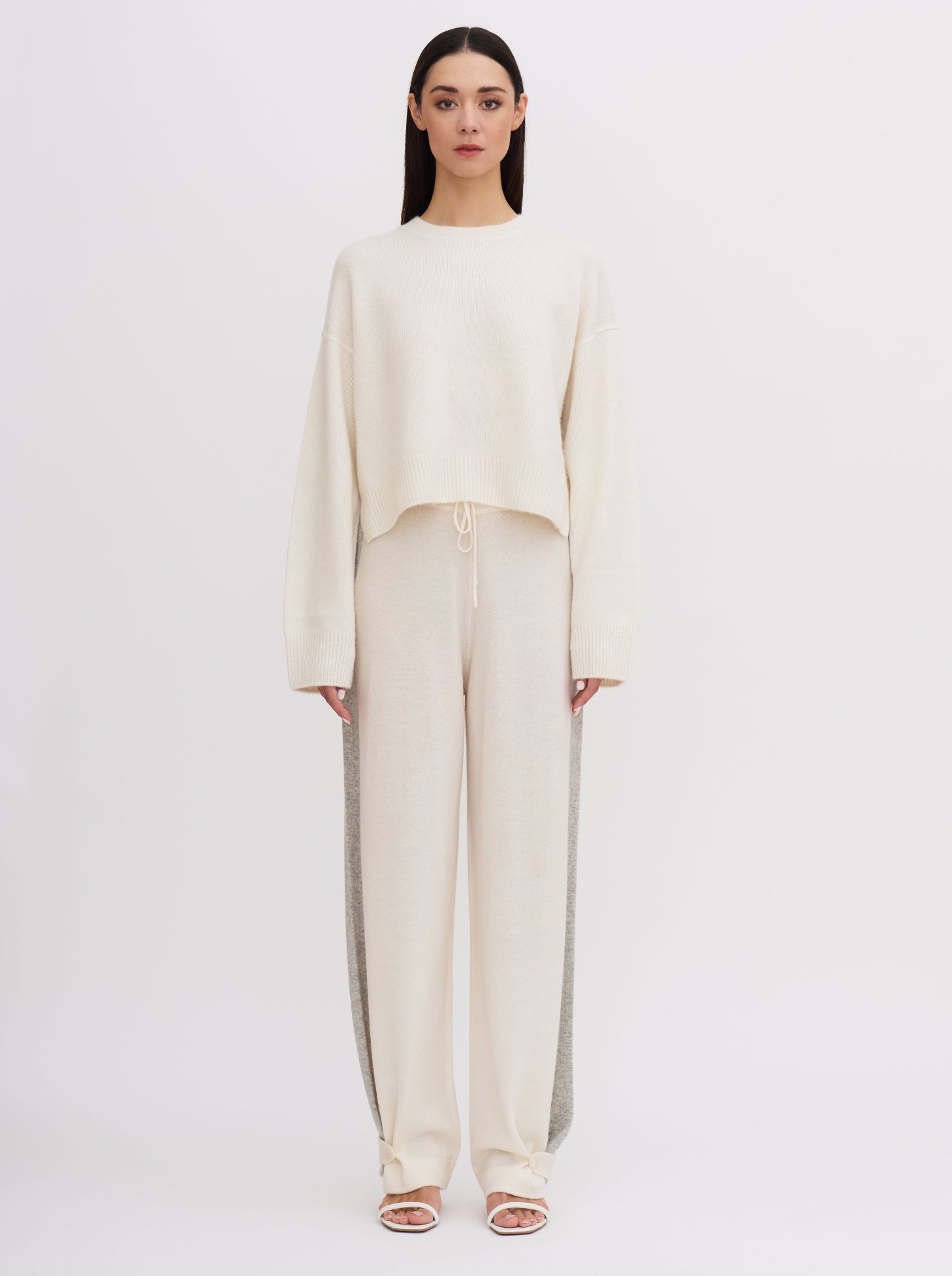 Front view of model wearing white wide leg pants with grey side stripes, front waist tie and buttoned ankle strap detail; worn with matching white cashmere jumper.