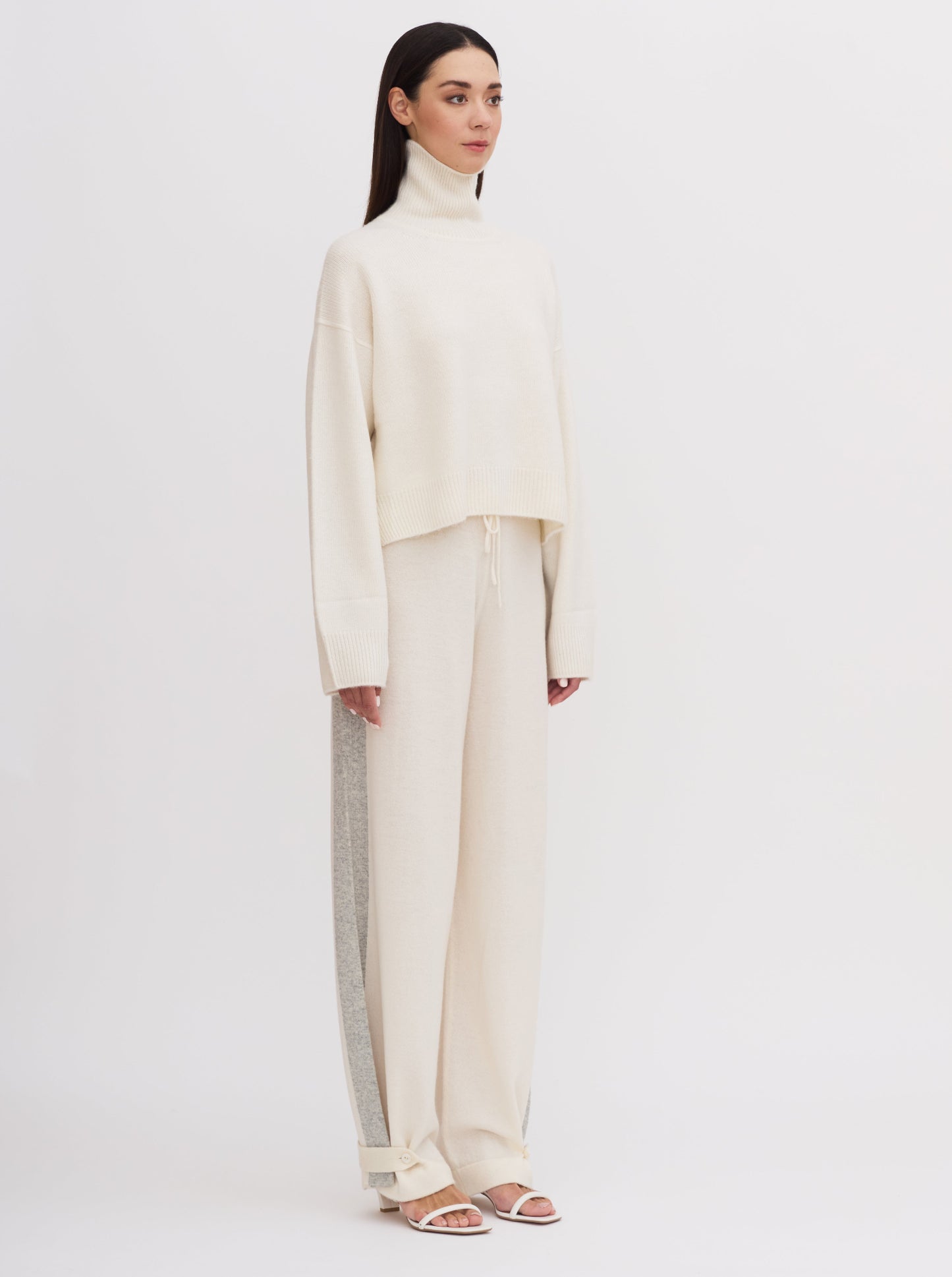 Model wearing white wide leg pants with grey side stripes, front waist tie and buttoned ankle strap detail; worn with white roll neck cashmere jumper.