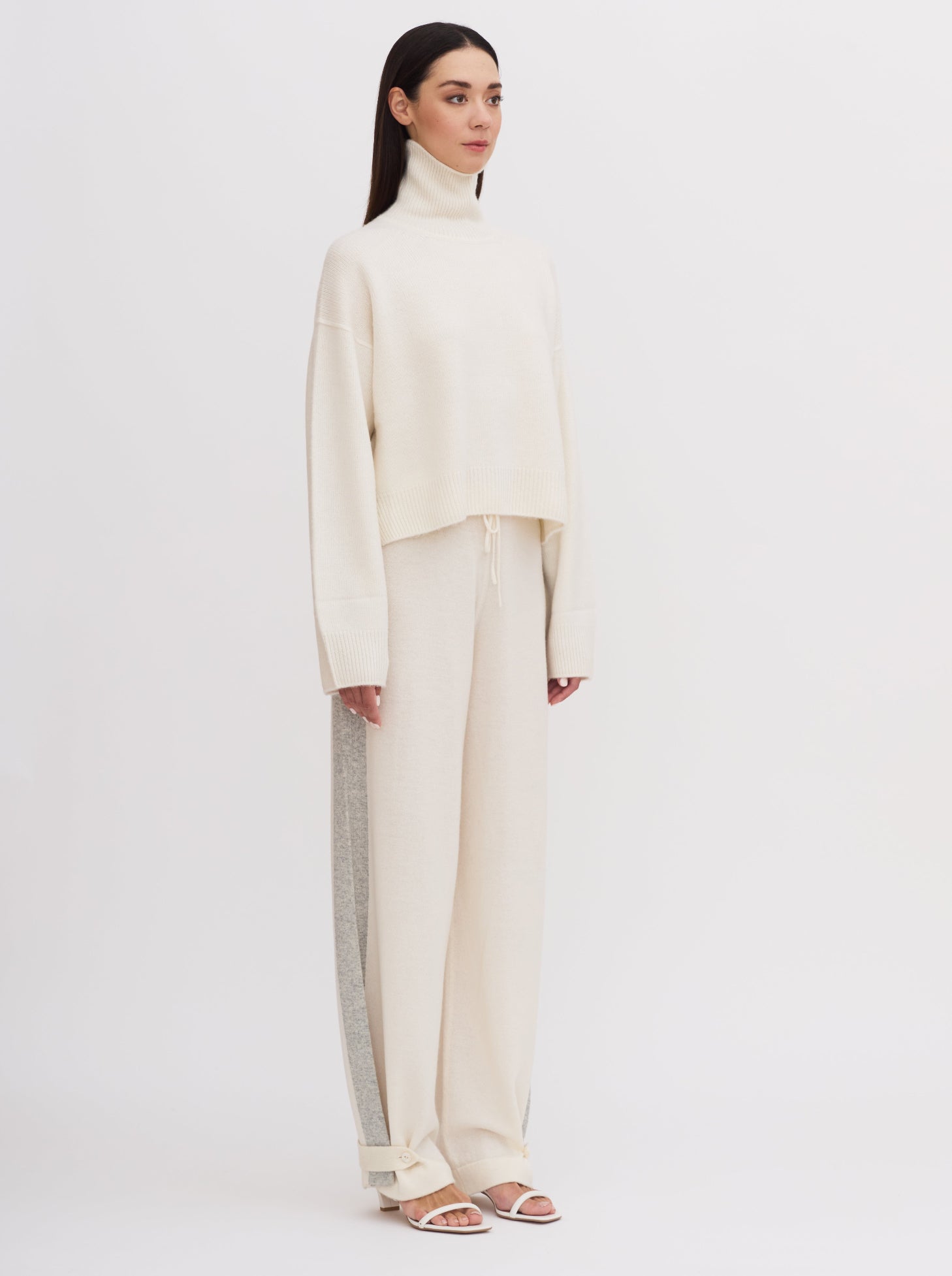 Model wearing white wide leg pants with grey side stripes, front waist tie and buttoned ankle strap detail; worn with white roll neck cashmere jumper.