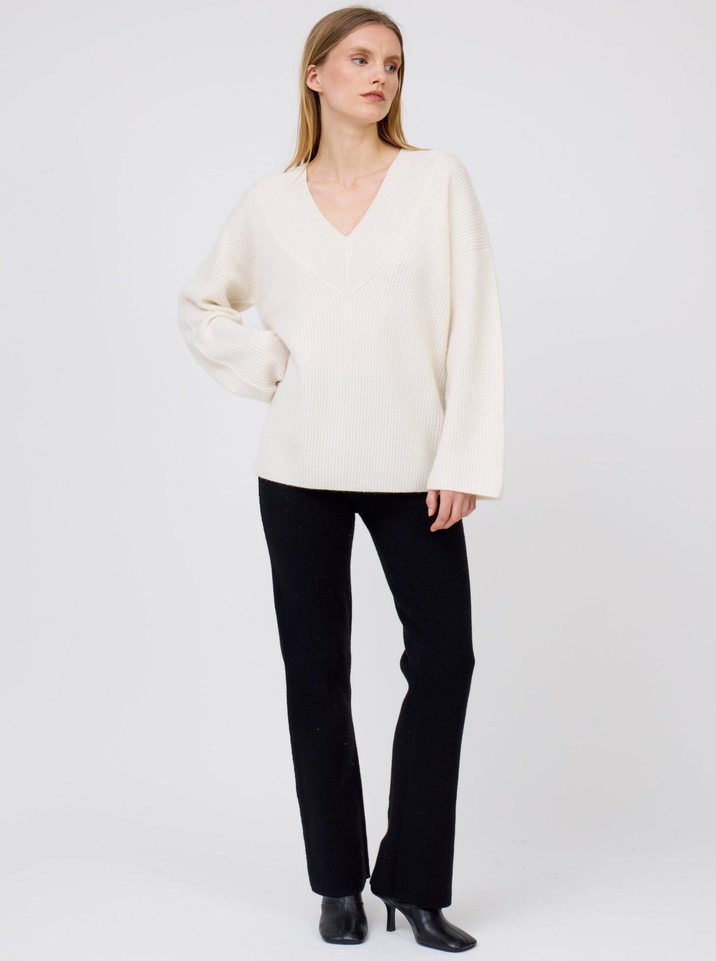 Model wears white deep V-neck ribbed knit cashmere sweater with generous long  sleeves; worn with black suit pants and black boots.