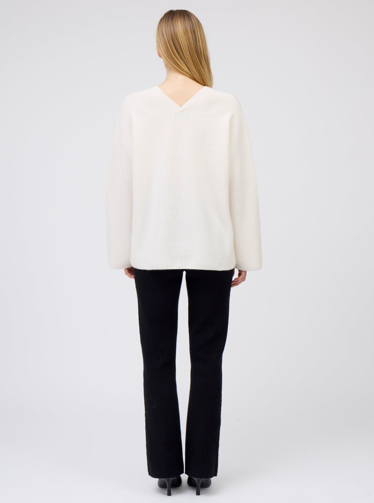 Back view of model wearing creamy white V-neck ribbed knit cashmere sweater with black tailored pants.