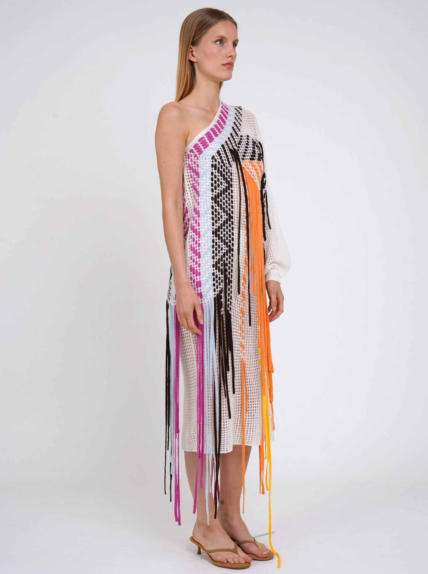 side view white light weight cashmere knit one shoulder shift dress with asymmetric neckline, decorated with woven coloured fringes which fall to the ground.