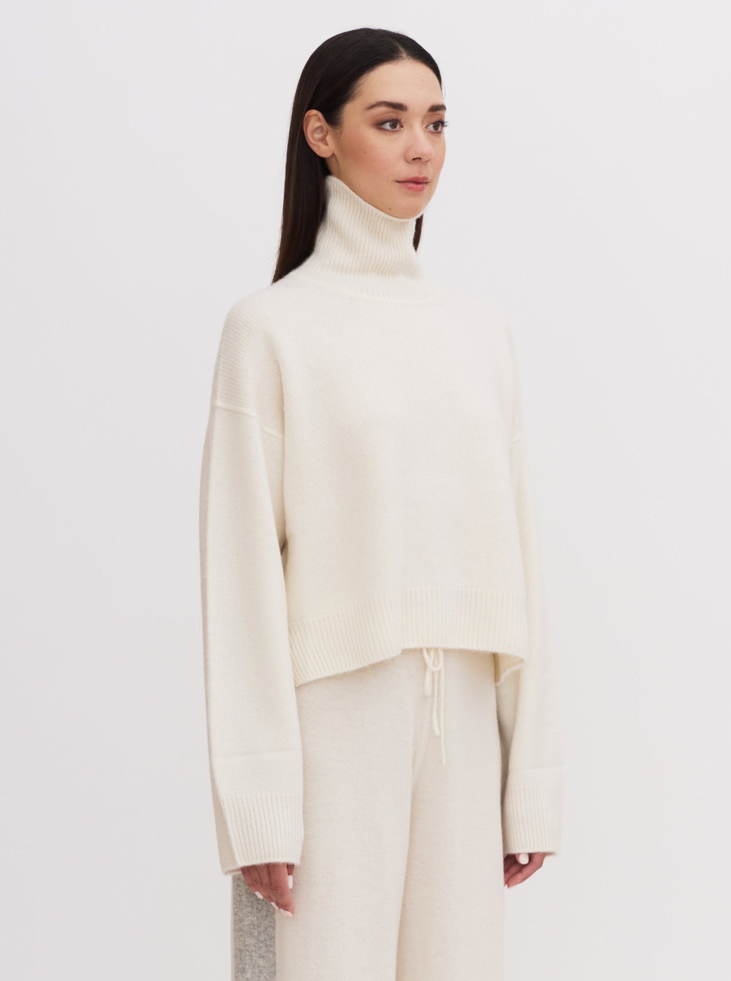 Side view of model wearing white cashmere, cropped length, roll neck sweater with wide sleeves and white wide leg pants.