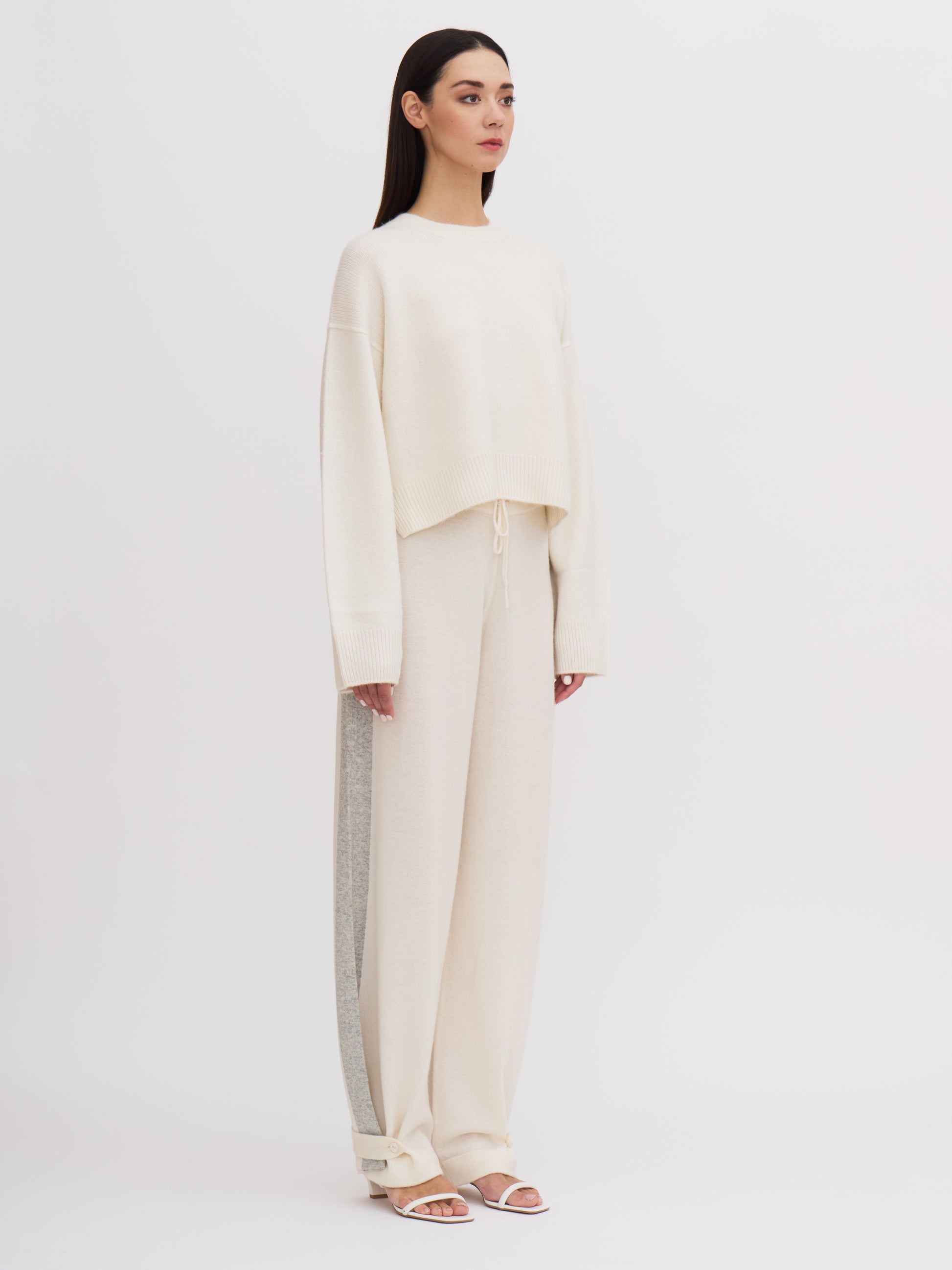 Side view of model wearing white cropped length round neck cashmere jumper and white wide leg pants with grey side stripe.