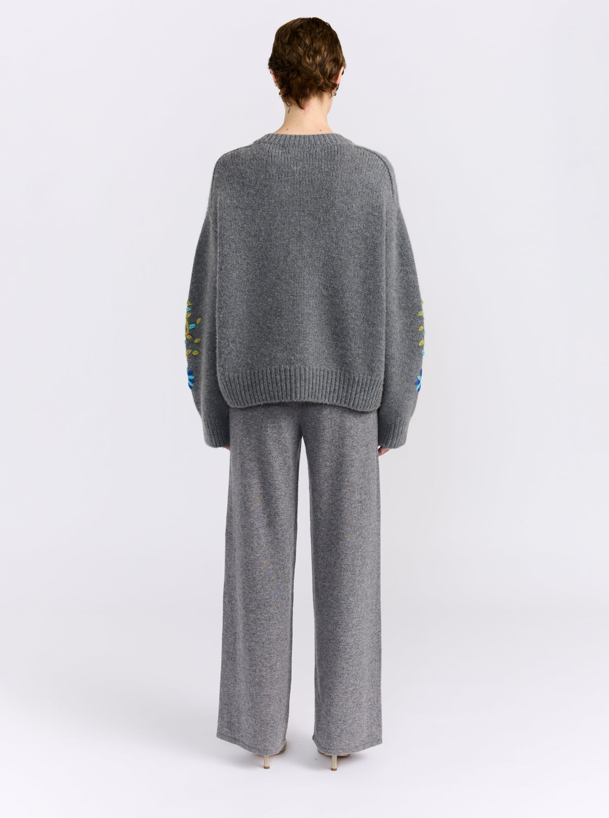 Back view model wearing a grey chunky knit oversized cardigan with and grey cashmere pants in a white studio.