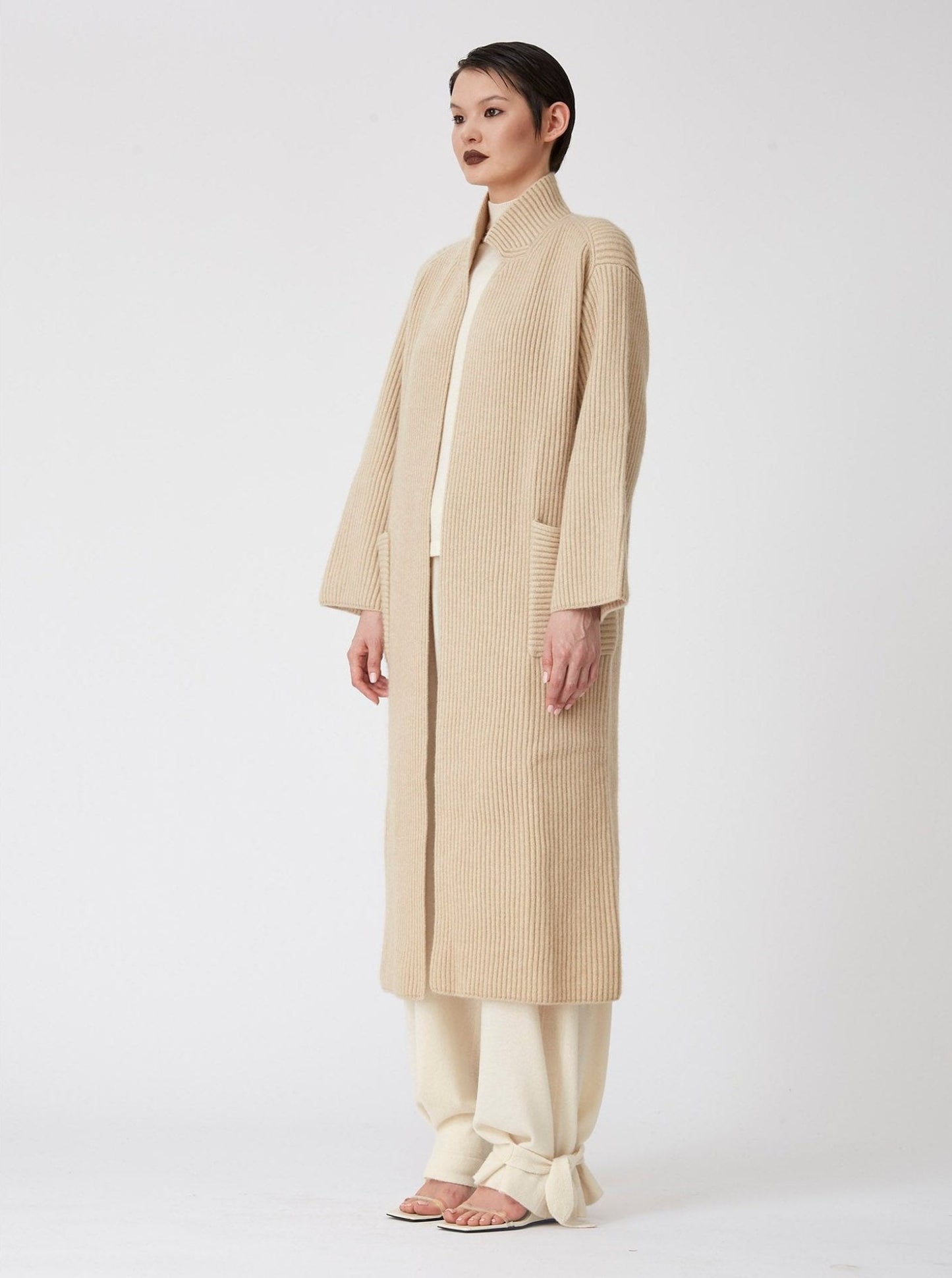 Side view of model wearing a beige Midi cardigan coat in ribbed cashmere knit. Straight cut with a relaxed fit, deep patch pockets and generous sleeves which can be rolled back. 
