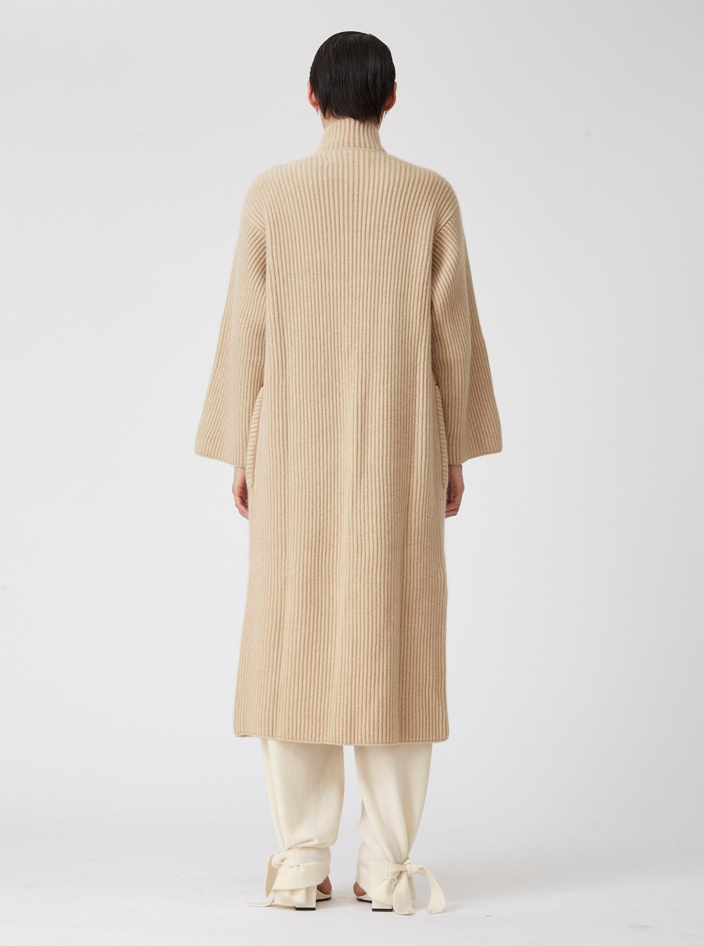 Back view of model wearing a beige midi cardigan coat in ribbed cashmere knit. Straight cut with a relaxed fit, deep patch pockets and generous sleeves which can be rolled back. 