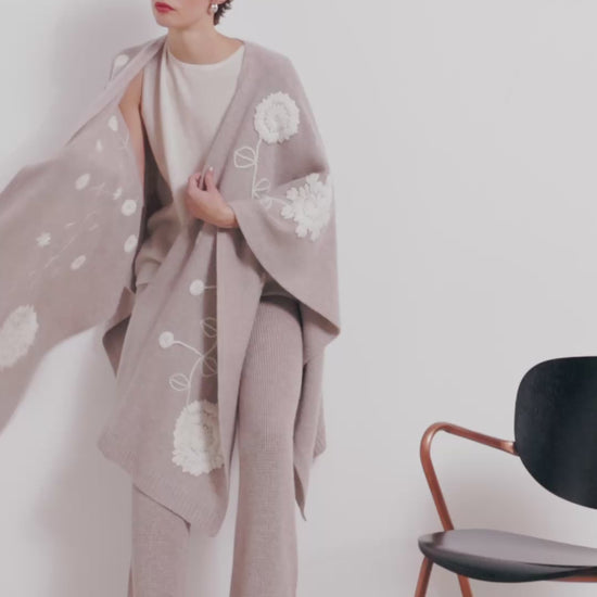 video clip model model wears beige cashmere long shawl with large white embroidered flowers over white knit top and beige knit pants.