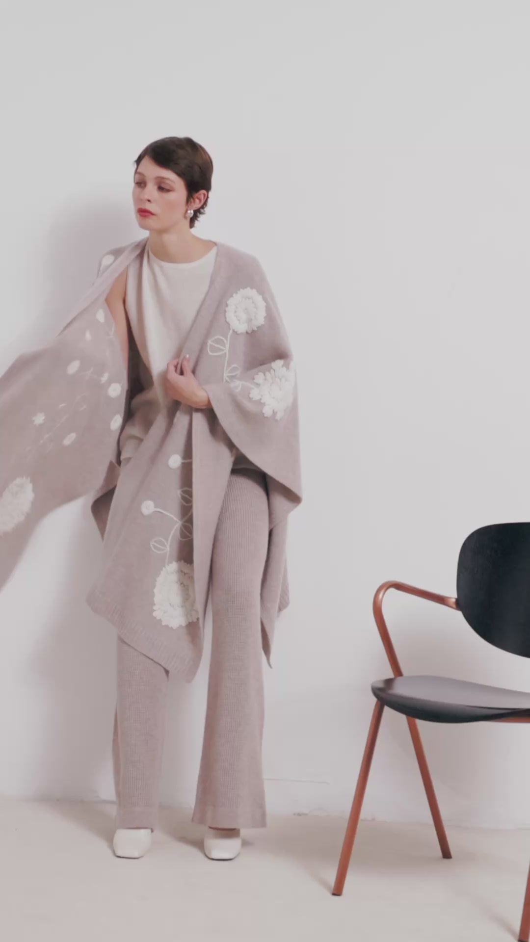 video clip model model wears beige cashmere long shawl with large white embroidered flowers over white knit top and beige knit pants.